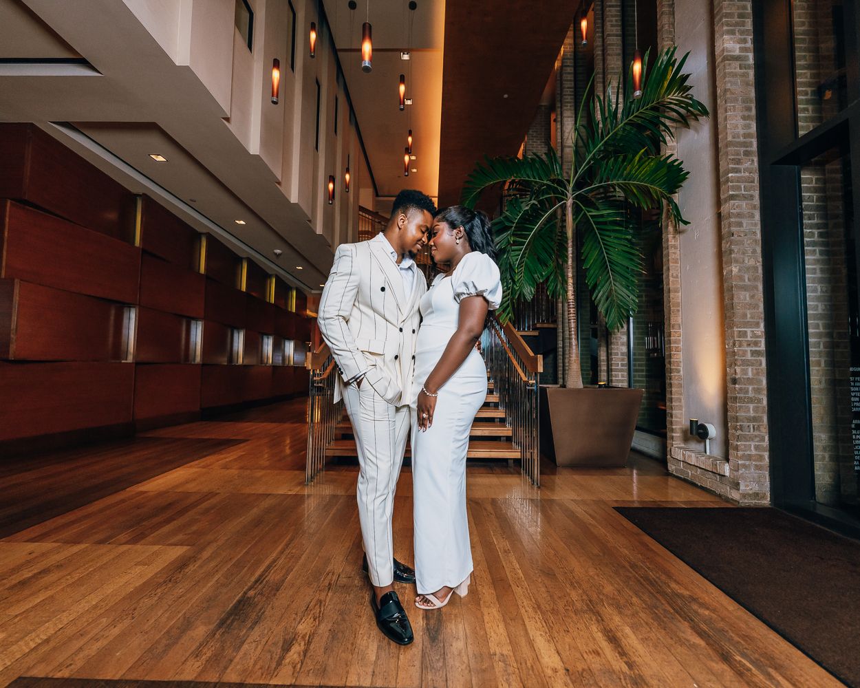 Couple in white outfits dance together on hardwood floors with indoor plants in background.