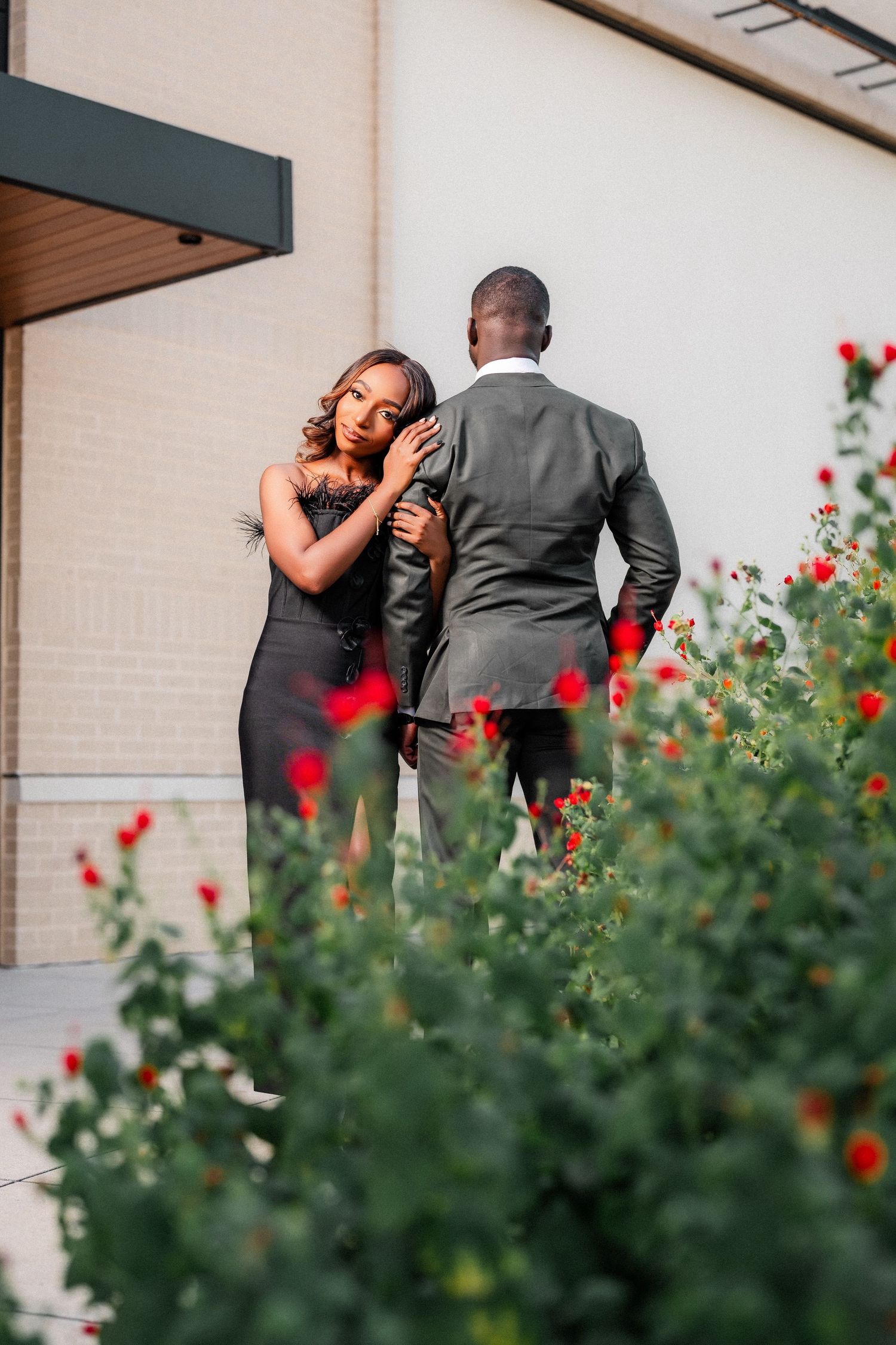 Romantic couple embracing near red flowers and a white building wall.