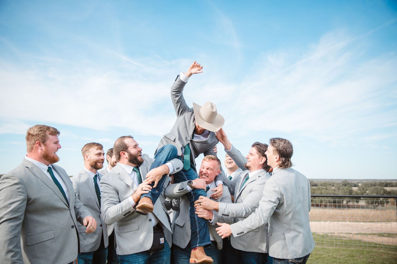 Groomsmen in gray suits celebrate outdoors against blue sky.