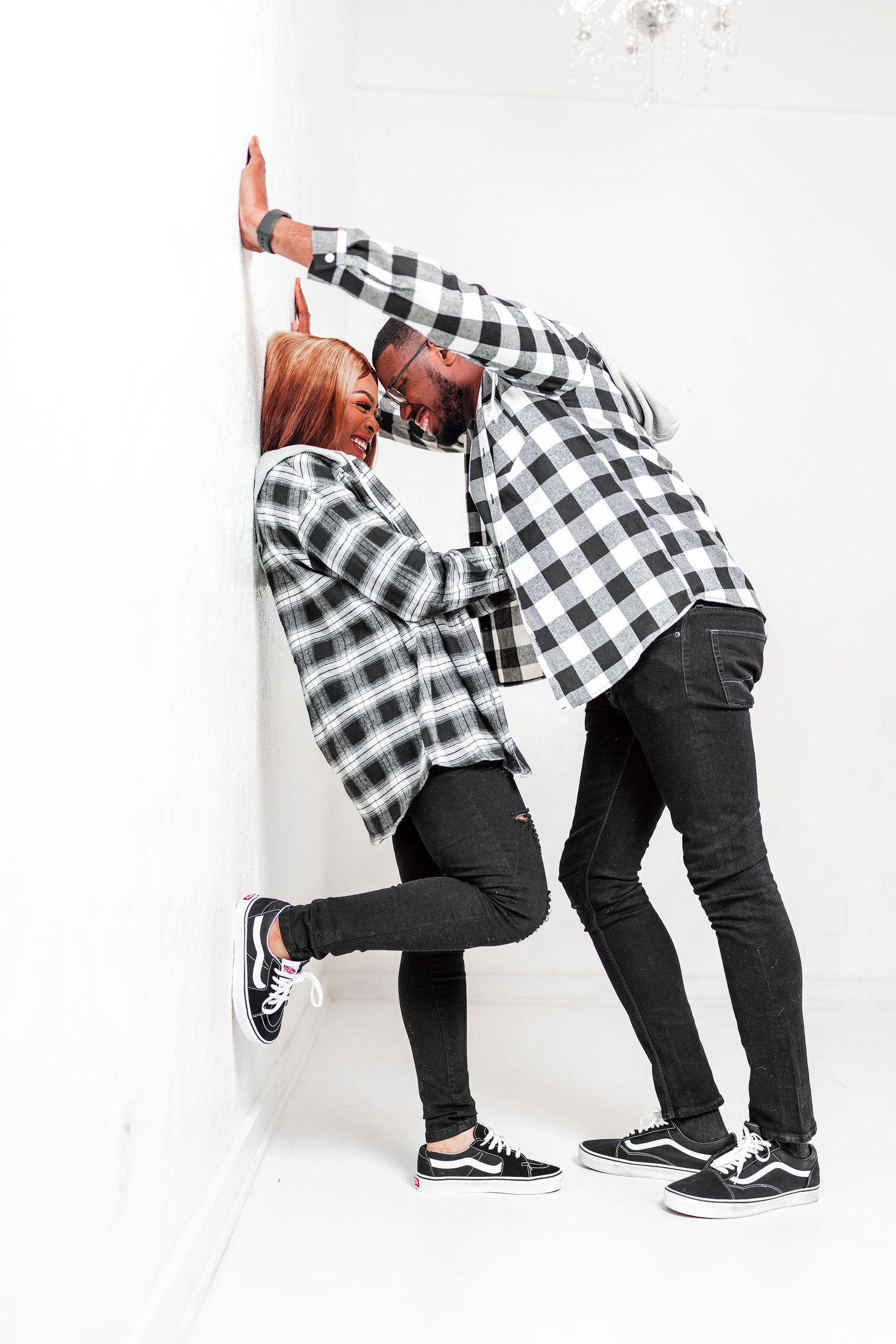 Two people in matching black and white plaid shirts and dark pants lean playfully against a white wall.
