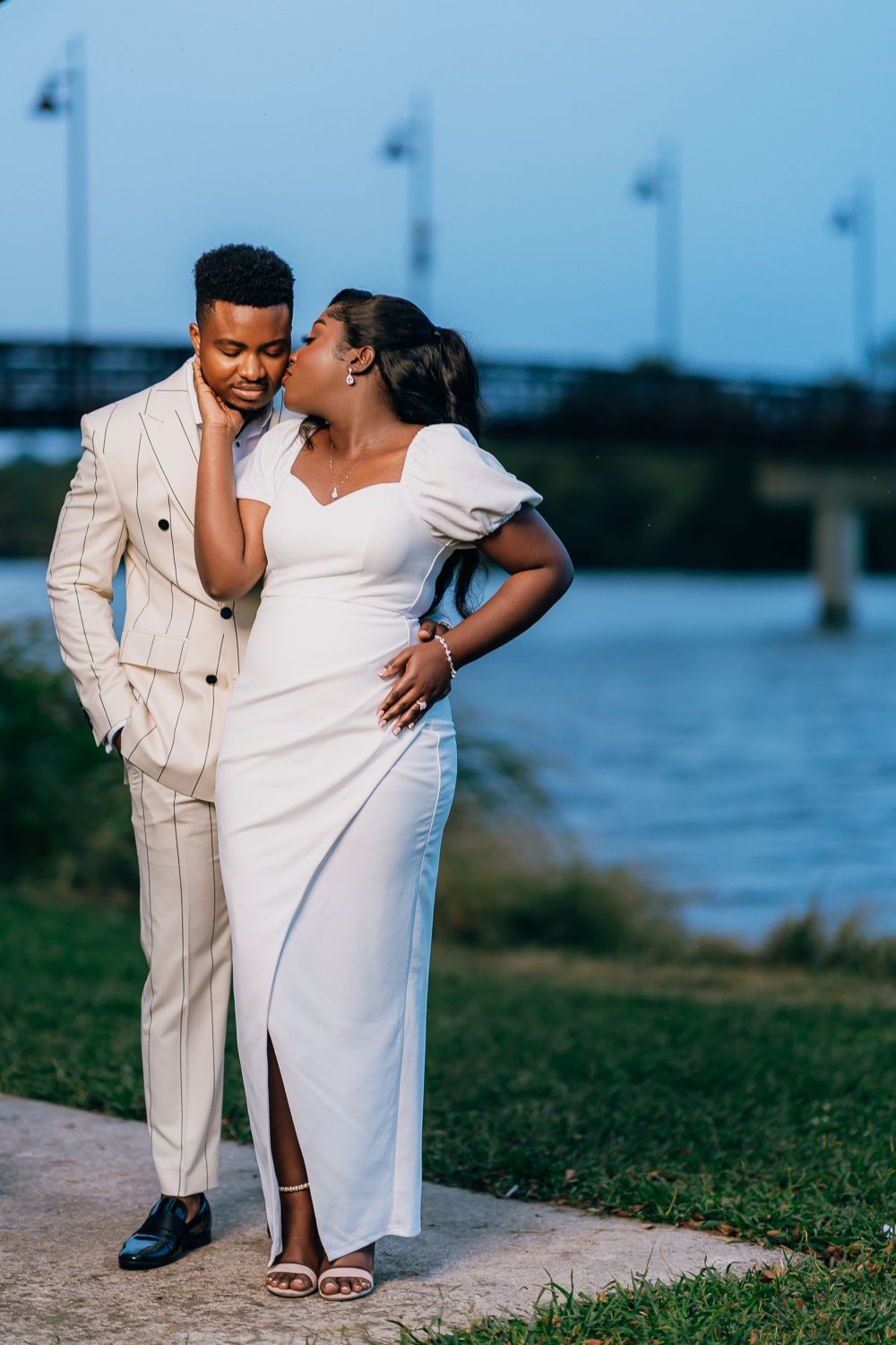 A romantic couple in elegant white outfits share an intimate moment by the waterfront at dusk.