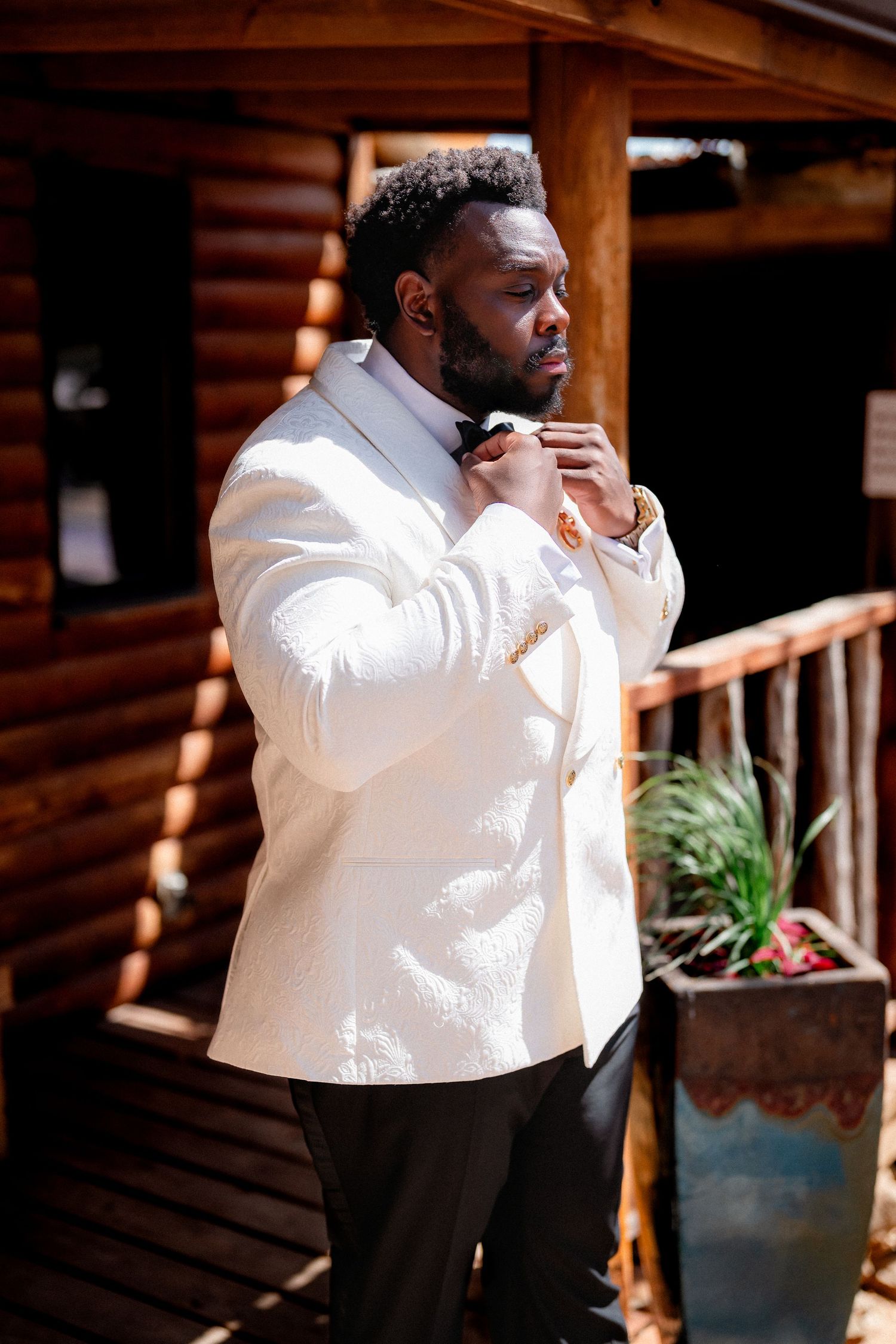 A person in a white tuxedo jacket adjusts their bowtie while standing on a rustic cabin porch.