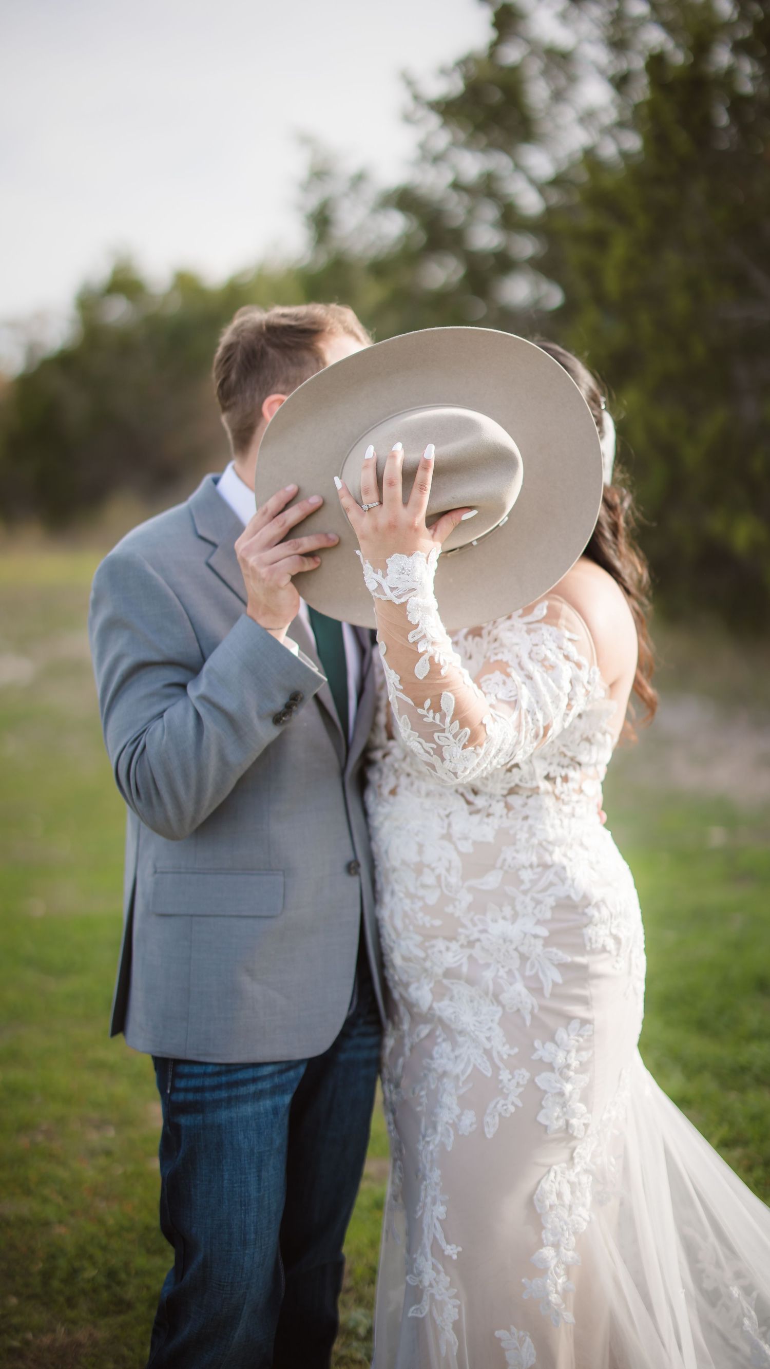 A couple shares a romantic moment behind a large beige cowboy hat during their outdoor wedding photoshoot.