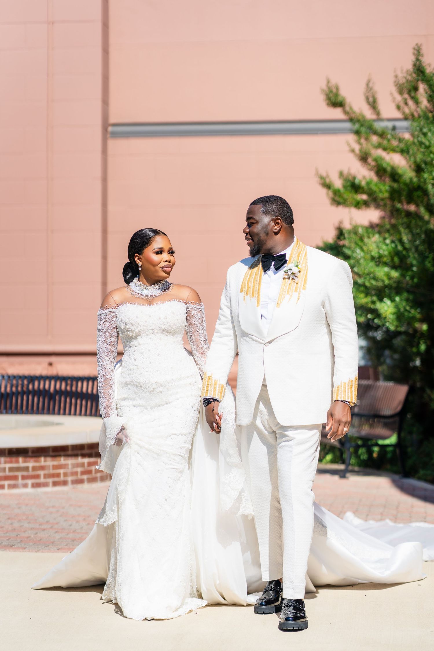 A couple in white wedding attire sits and poses together on outdoor steps with a pink wall backdrop.
