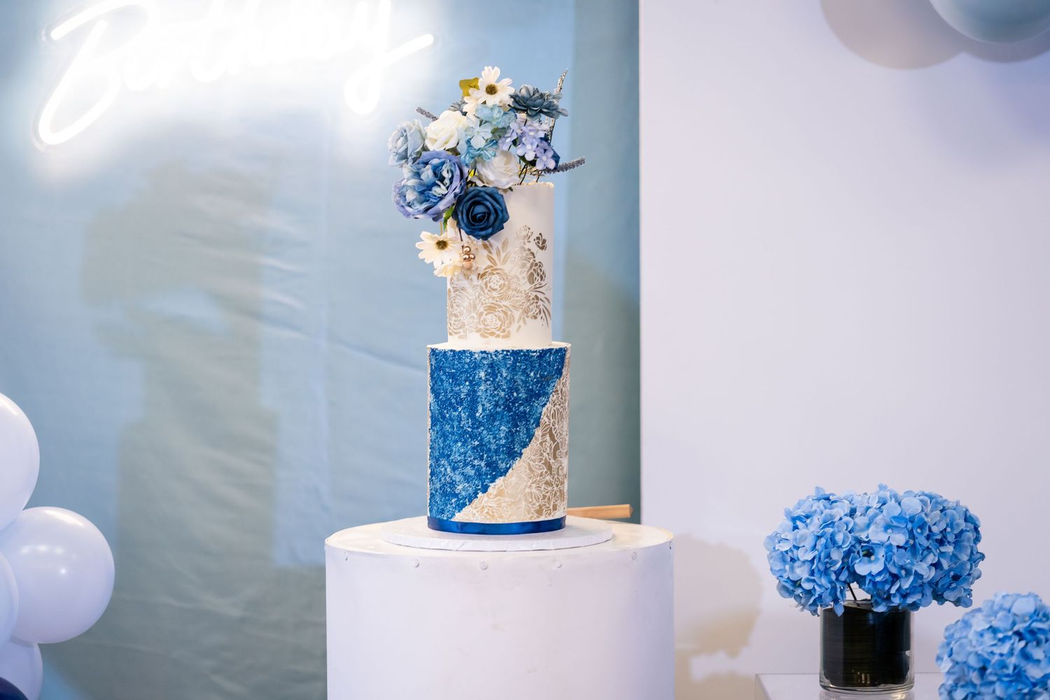 Elegant white and blue wedding cake decorated with hydrangeas and delicate floral details on a white pedestal.