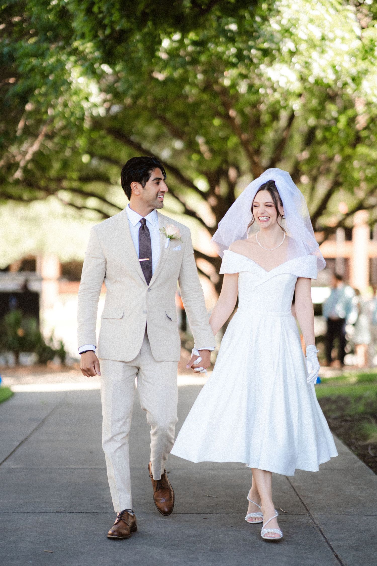 A wedding couple walks together on a tree-lined path, the bride in white and groom in beige.