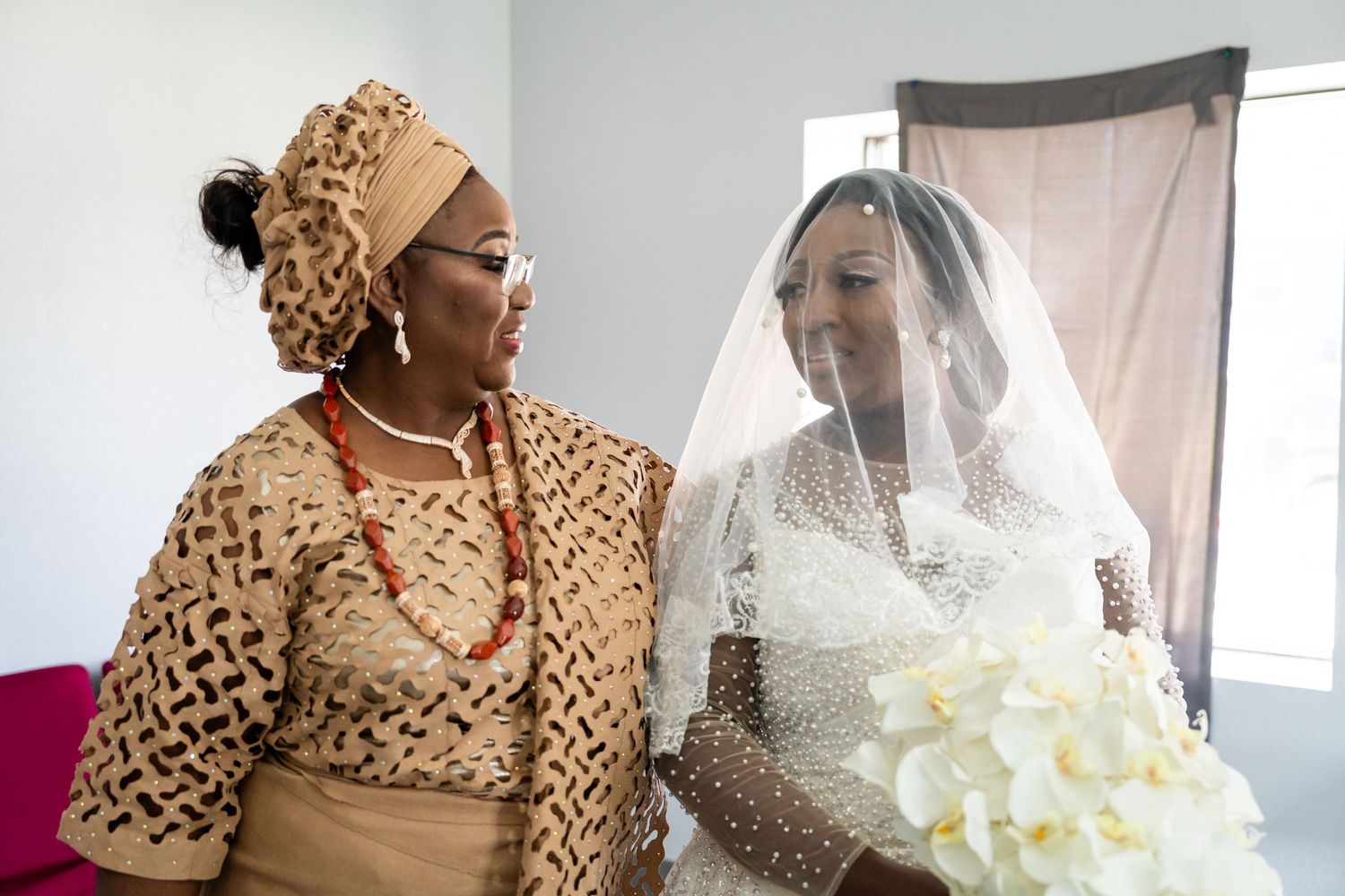 A bride in a white wedding dress and veil sits with a companion wearing traditional African attire and headwrap.