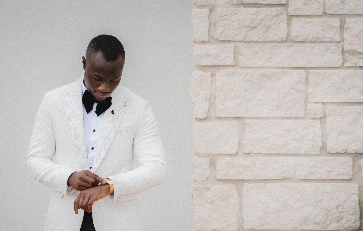 Person in white tuxedo jacket and bow tie checking time on watch against stone wall background.