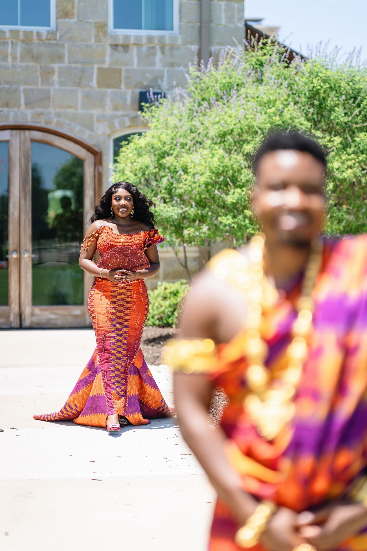 A person wearing a vibrant orange and red patterned dress poses outdoors against a stone building backdrop.
