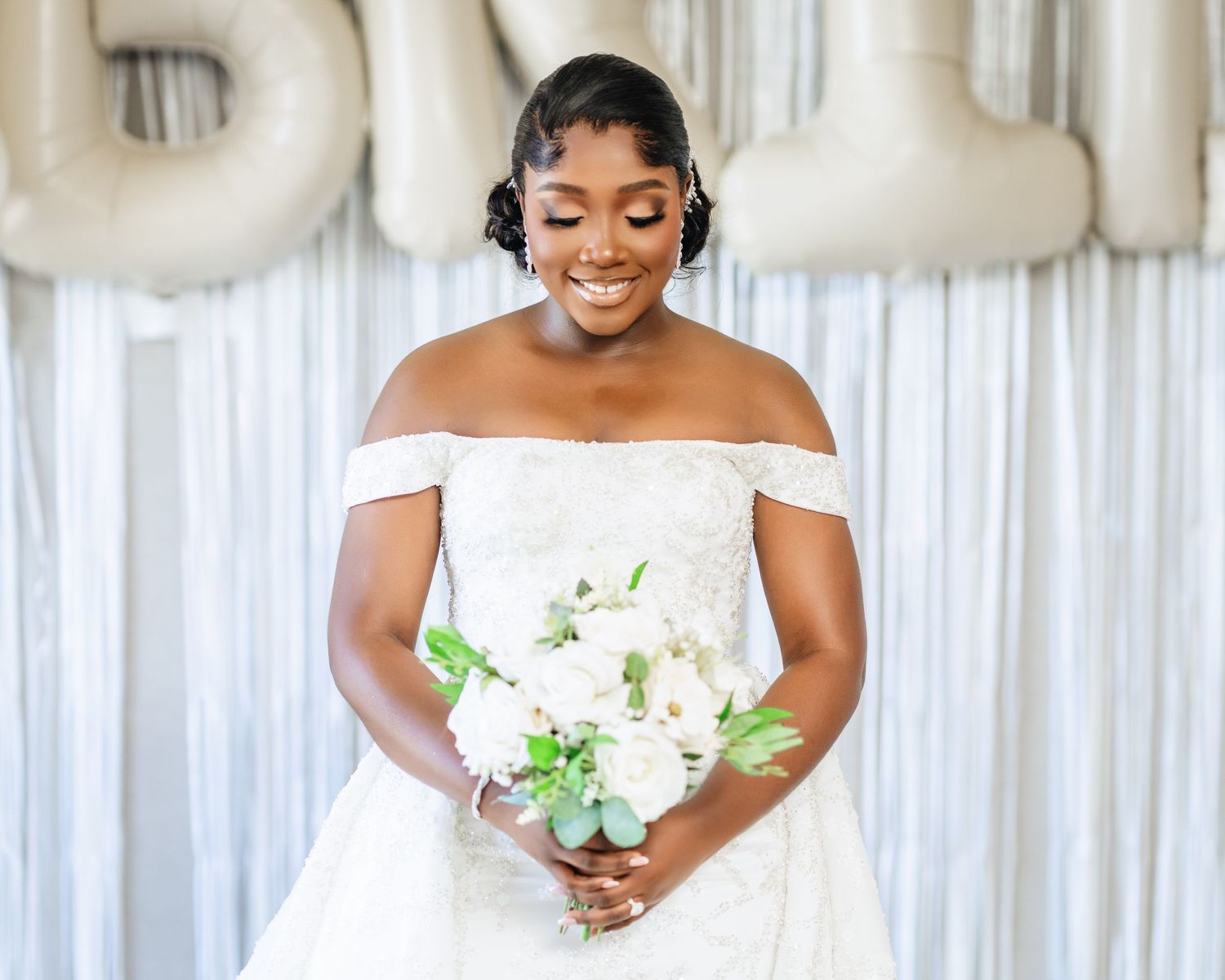A bride in a strapless white gown holds a white floral bouquet against a white draped backdrop.