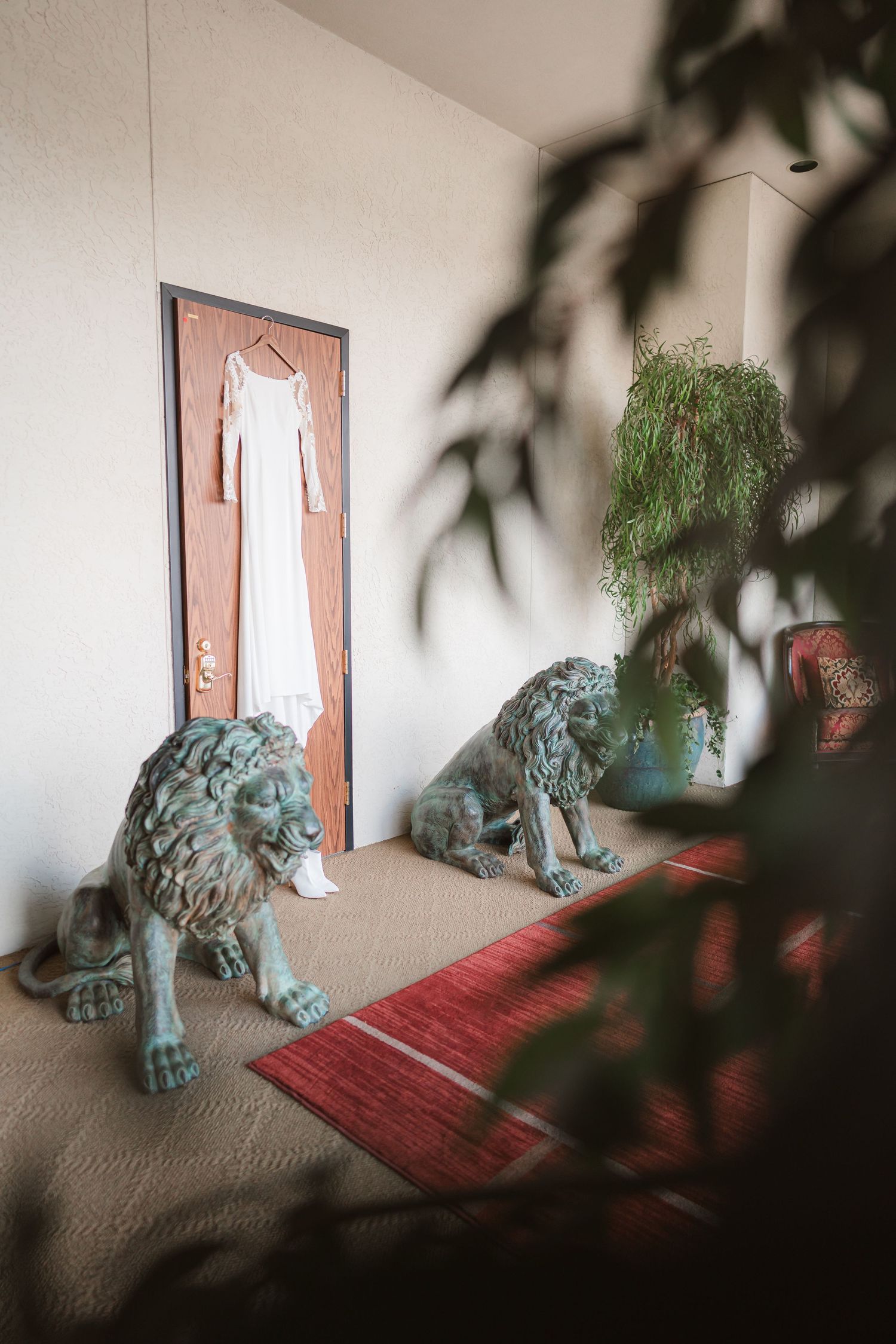 A white wedding dress hangs on a door with decorative blue lion statues and a red rug in the foreground.