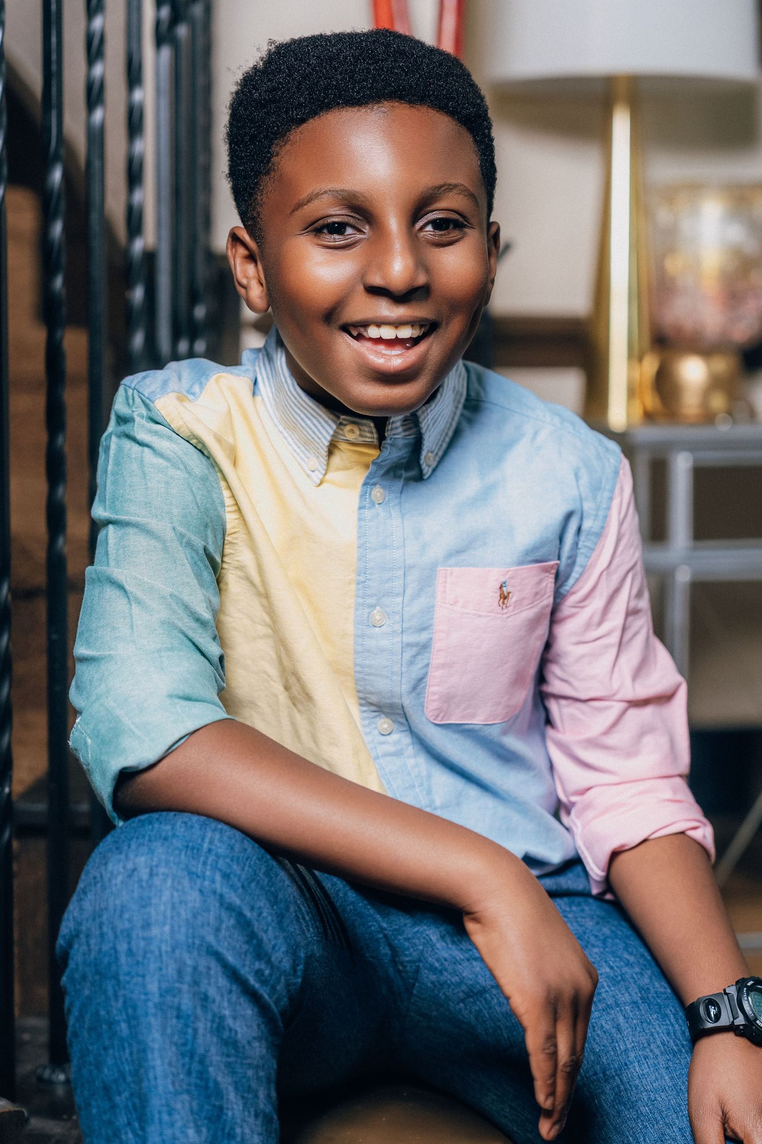 A young child wearing a color block pastel shirt and jeans poses while sitting and smiling in a casual indoor setting.