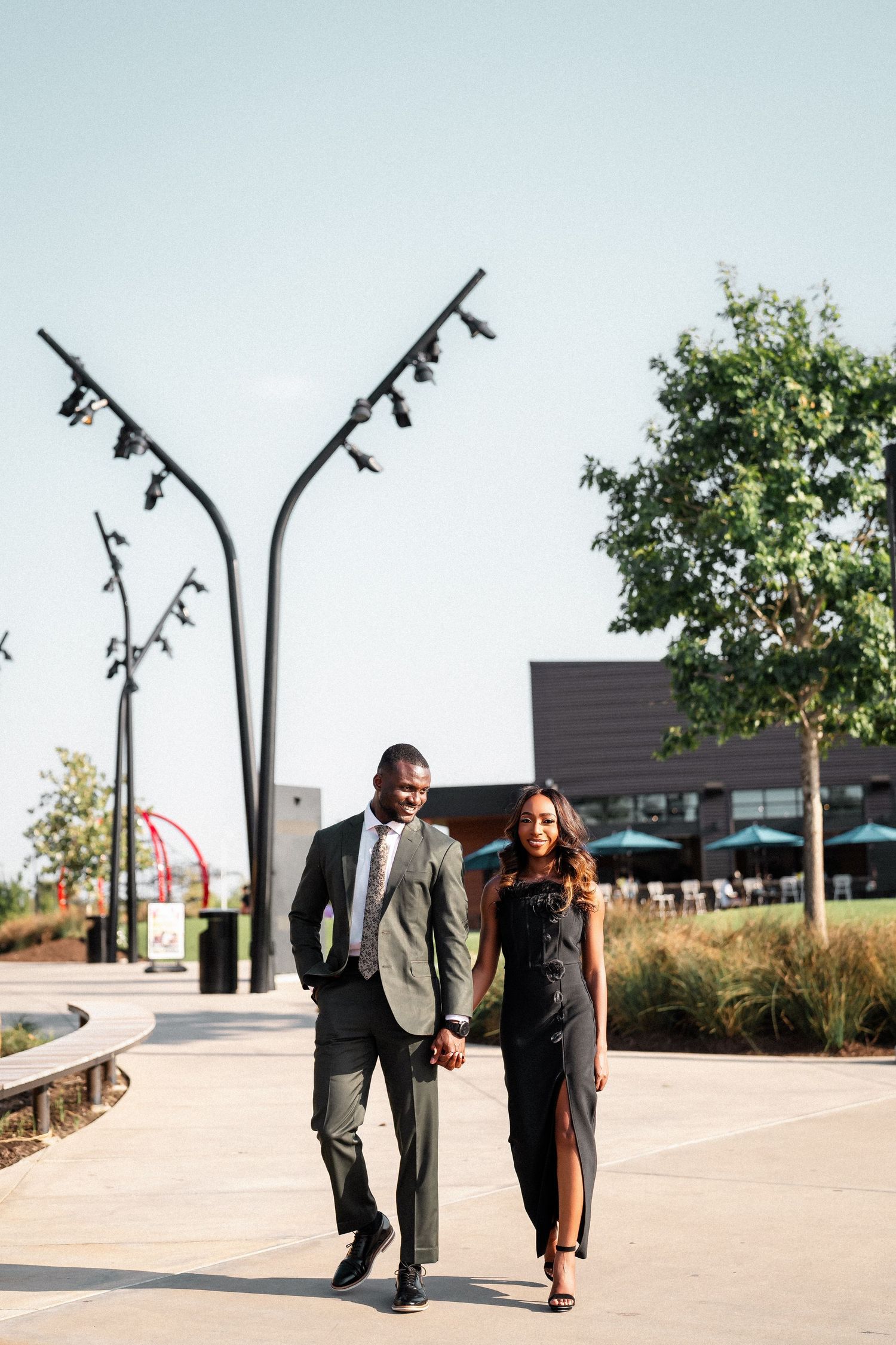 A couple walks together on a paved path with street lamps and landscaping in an urban setting.