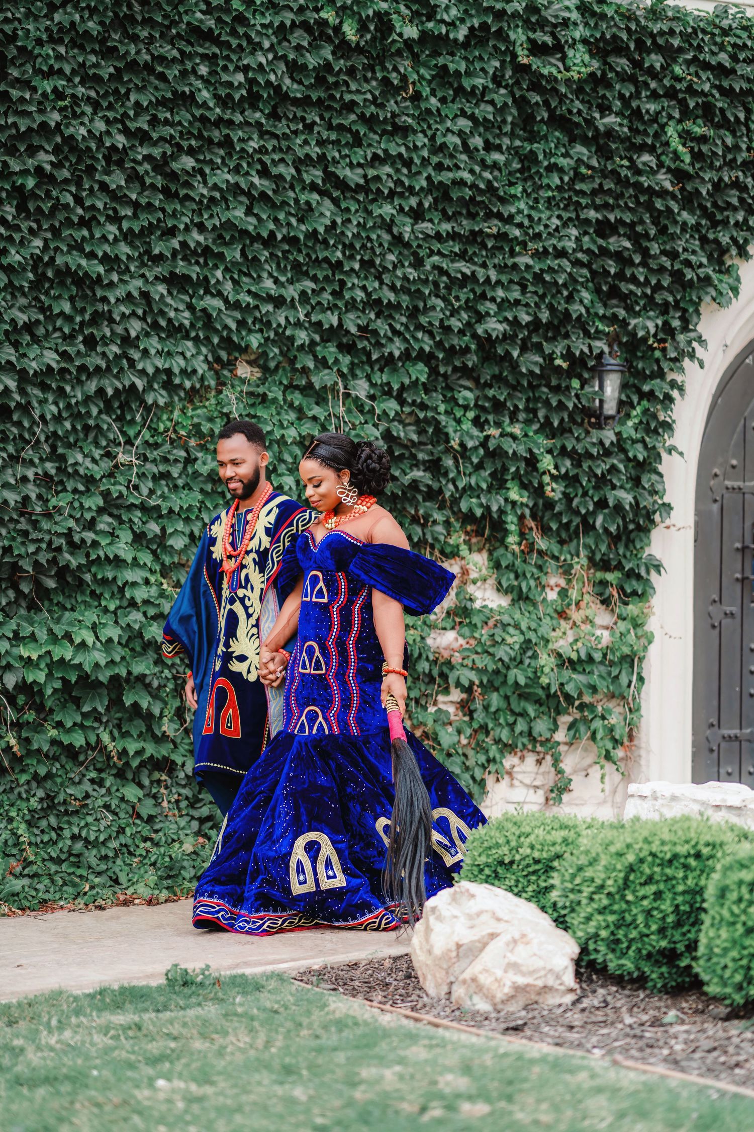A couple in vibrant blue traditional African attire poses against an ivy-covered wall.