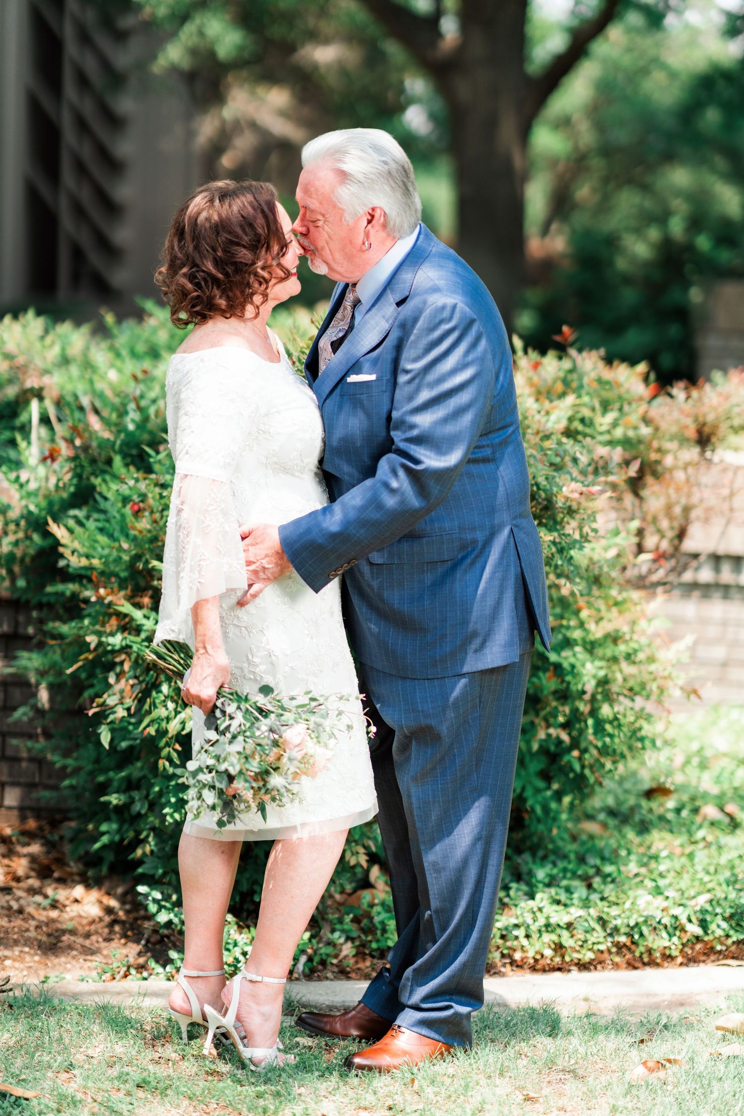 A couple shares an intimate moment in a garden setting, with the woman in white and man in blue suit.