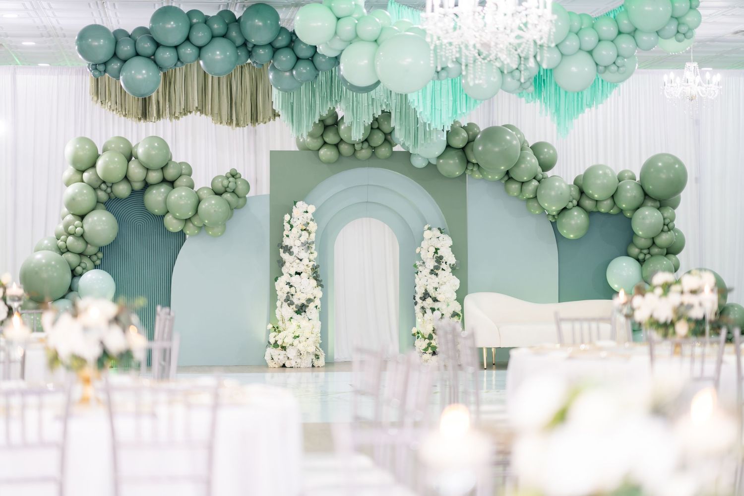 Elegant wedding reception decor with mint green balloon garlands and white floral arrangements against sage walls.