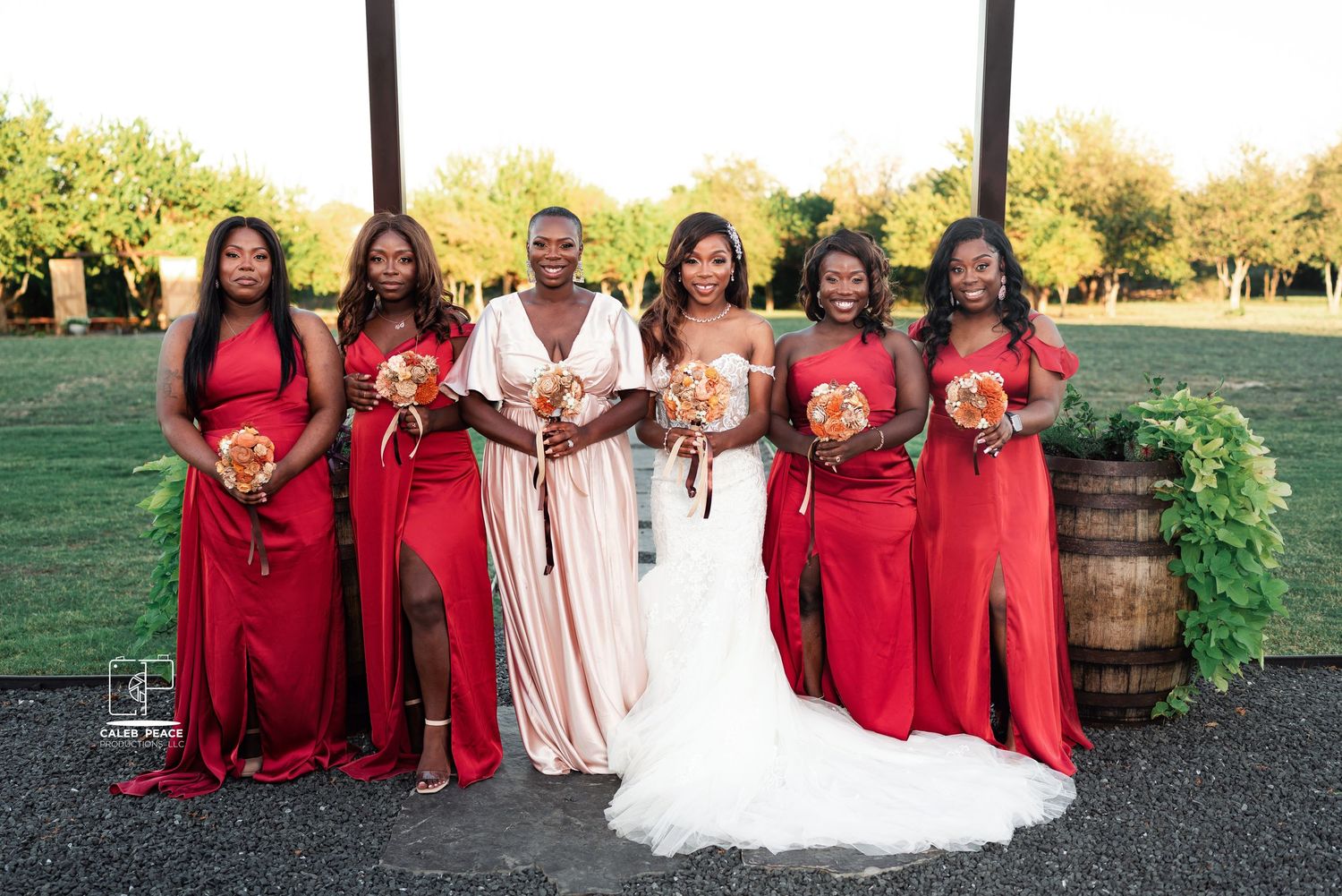 Wedding party in red dresses poses with couple against natural outdoor backdrop.