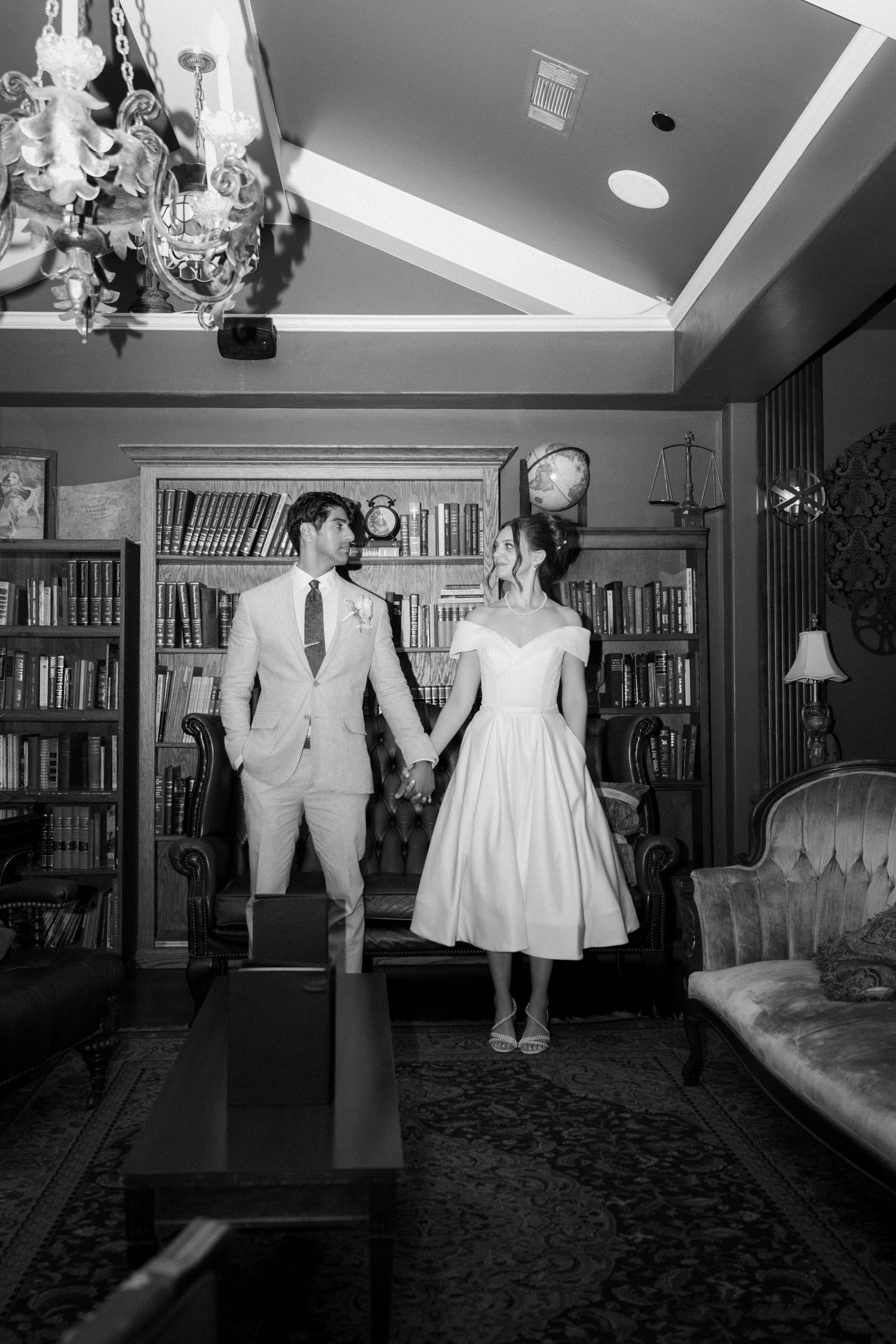 Black and white photo of newlyweds dancing together in a library setting with bookshelves in background.