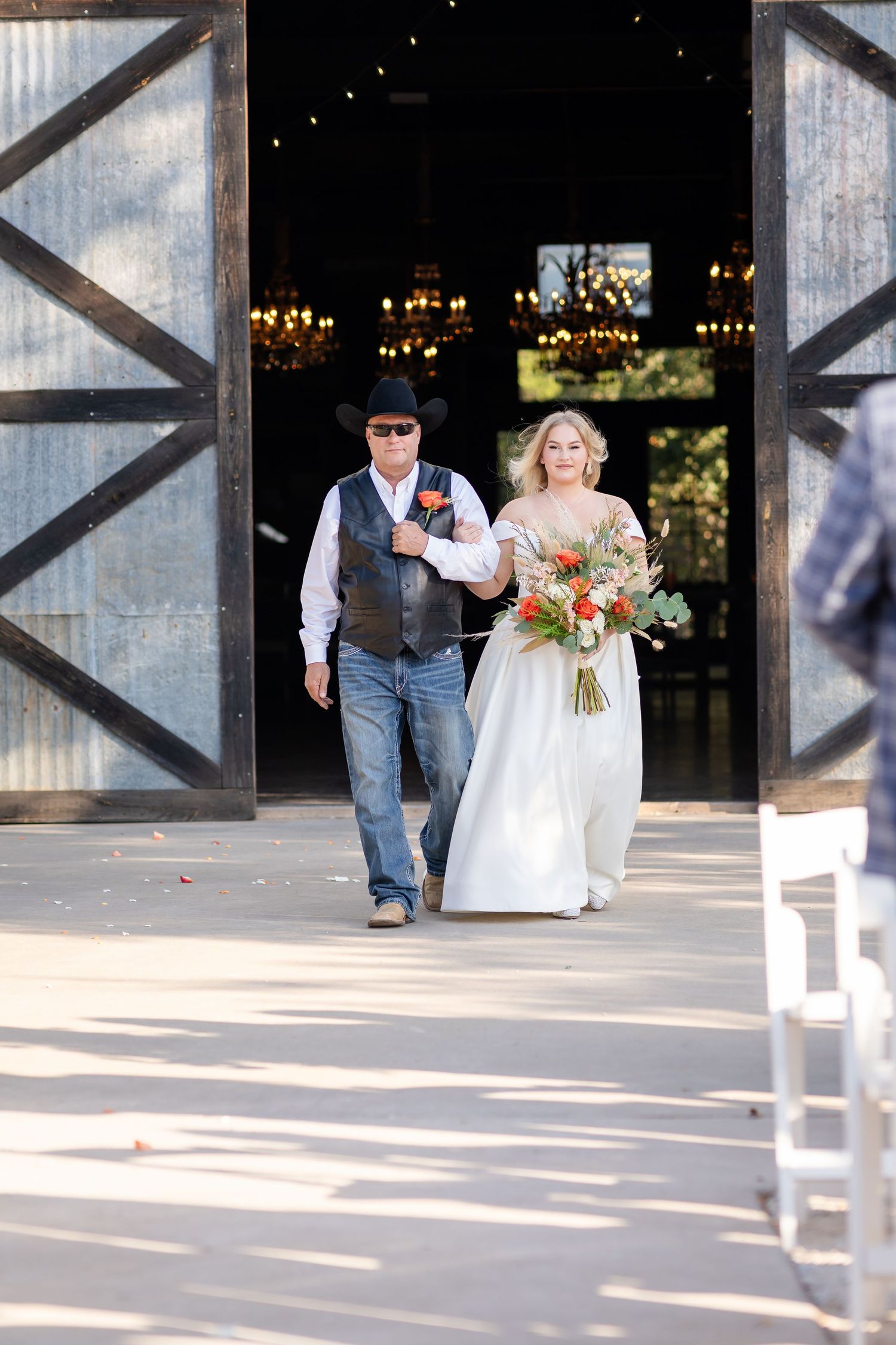 A couple walks down a modern venue's patio with white chairs and industrial architecture in the background.