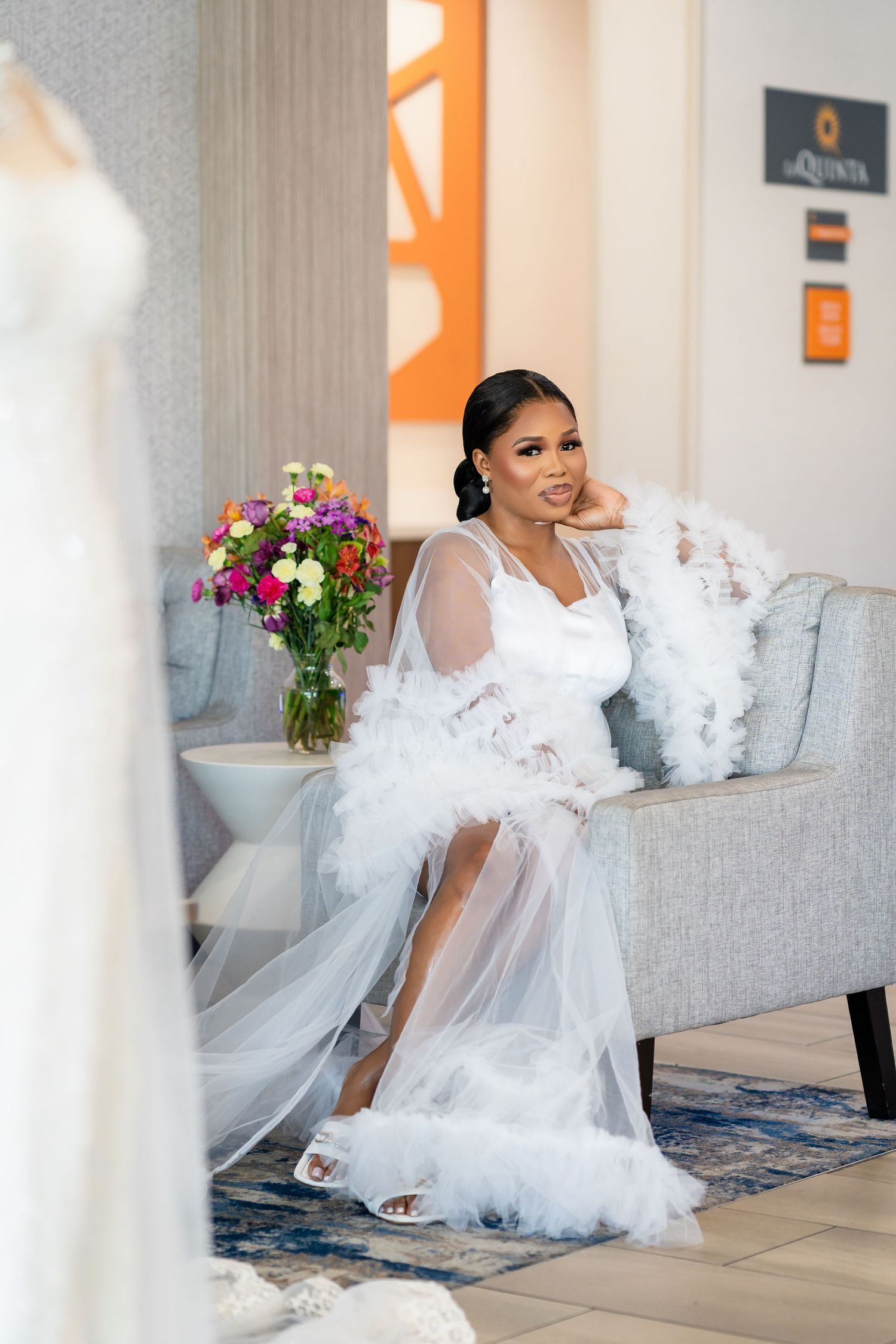 Bride in flowing white gown poses gracefully on grey couch with decorative pillows.