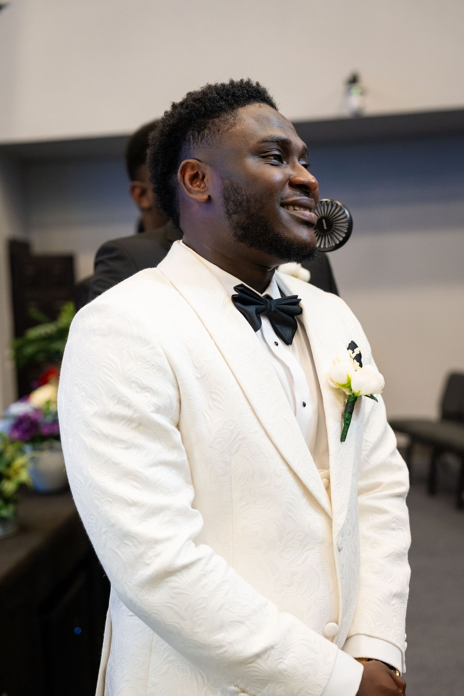 Groom in white tuxedo jacket with boutonniere smiling during wedding ceremony.