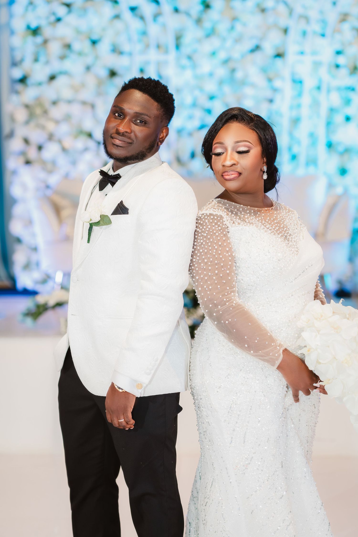 Newlyweds pose together in elegant white attire against a shimmering blue backdrop.