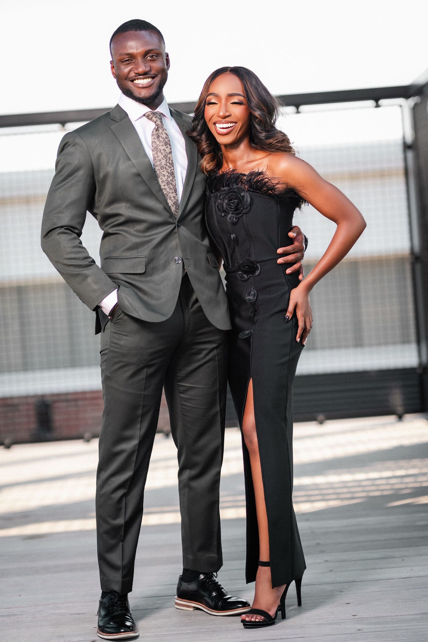 A stylishly dressed couple poses together outdoors, with him in a gray suit and her in an elegant black evening gown.