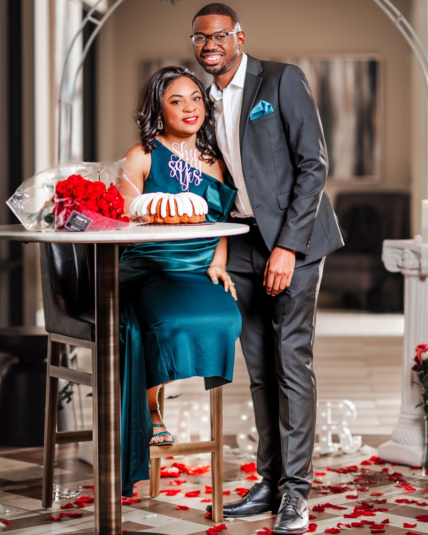 Romantic couple poses by elegant table with scattered red rose petals and teal decor.