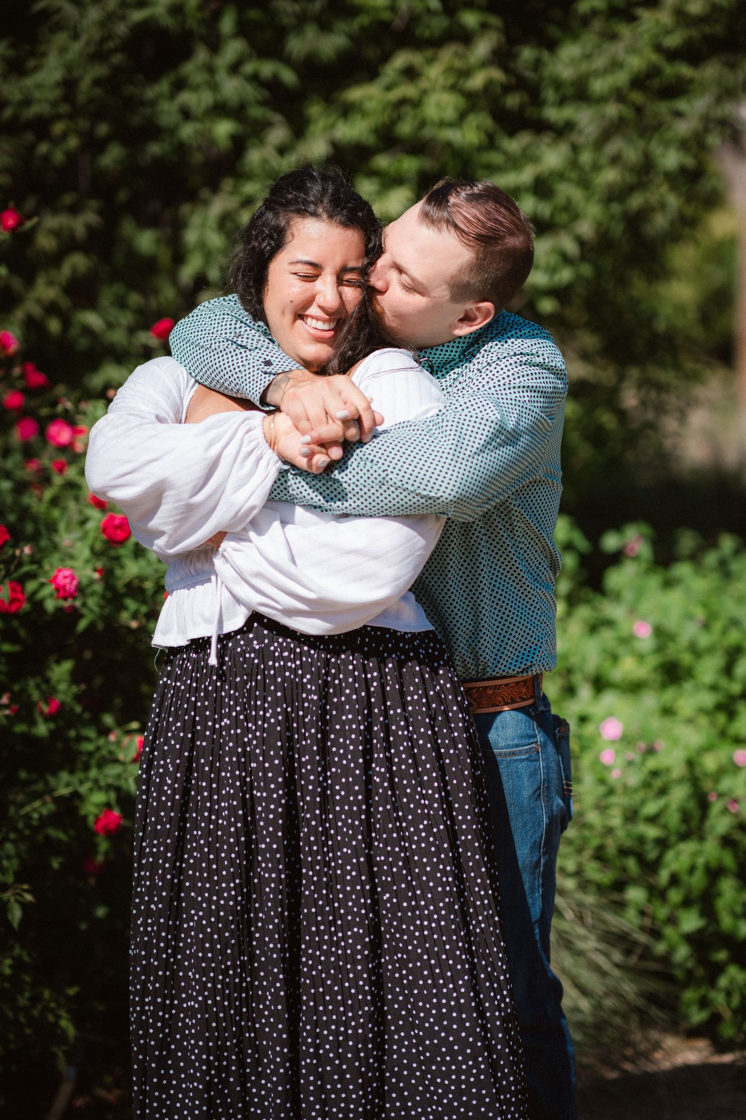 A romantic embrace in a garden setting with blooming flowers, featuring a polka dot skirt and blue checkered shirt.