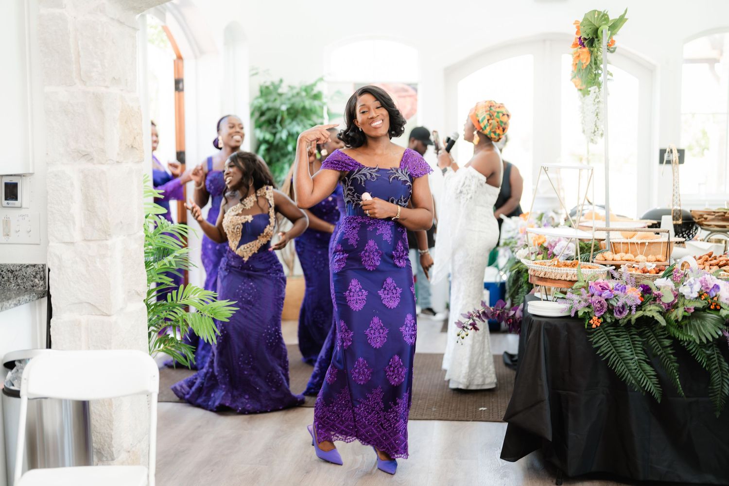 Wedding party in purple dresses and white gowns gather around a reception table with floral centerpieces.
