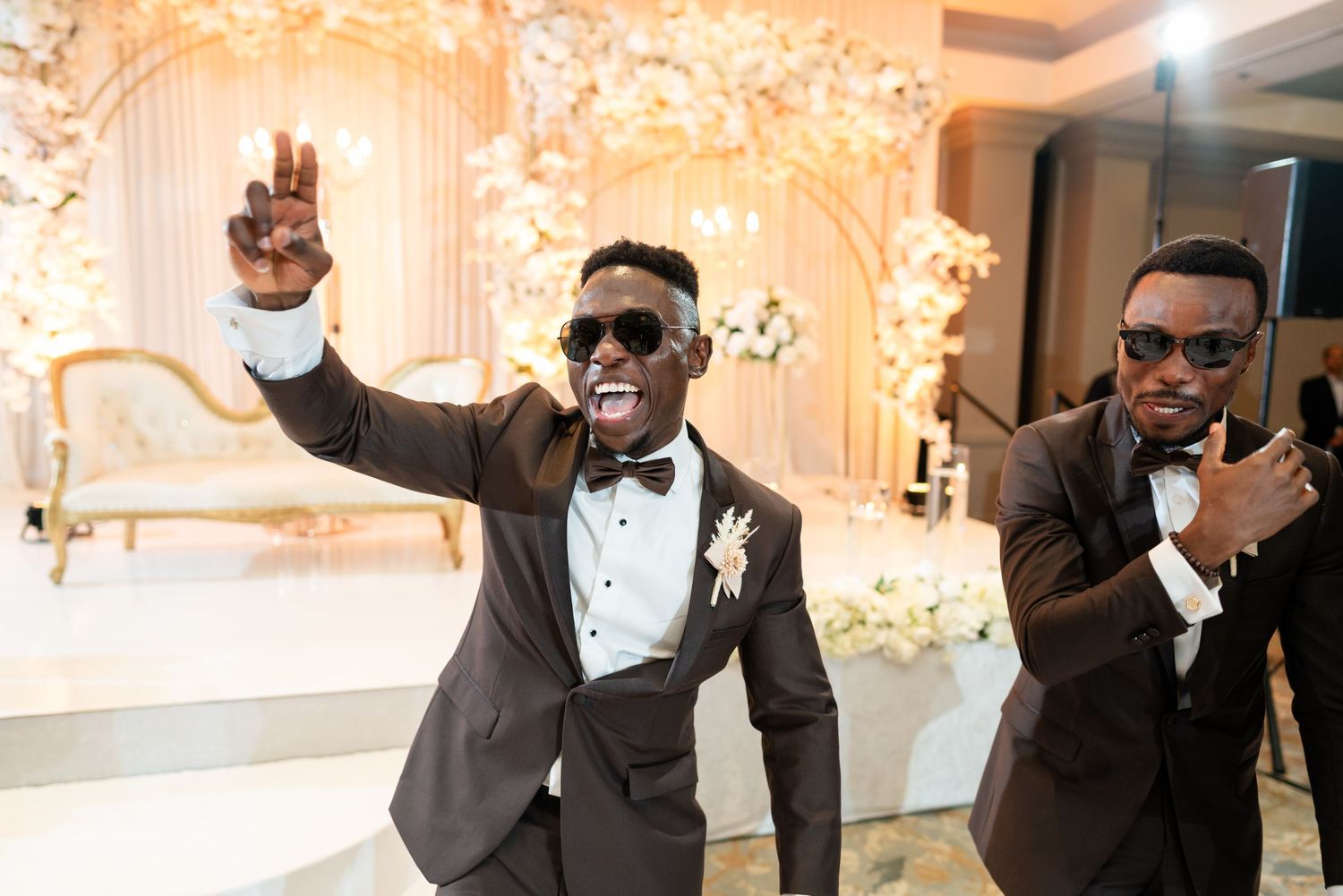 Groomsmen in black tuxedos and sunglasses pose playfully at a wedding celebration.