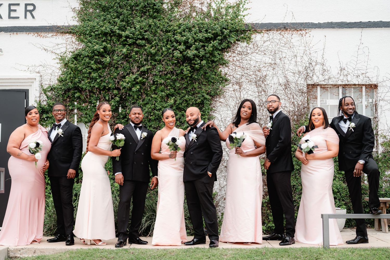 A wedding party in pink dresses and black suits lined up against a white building wall.