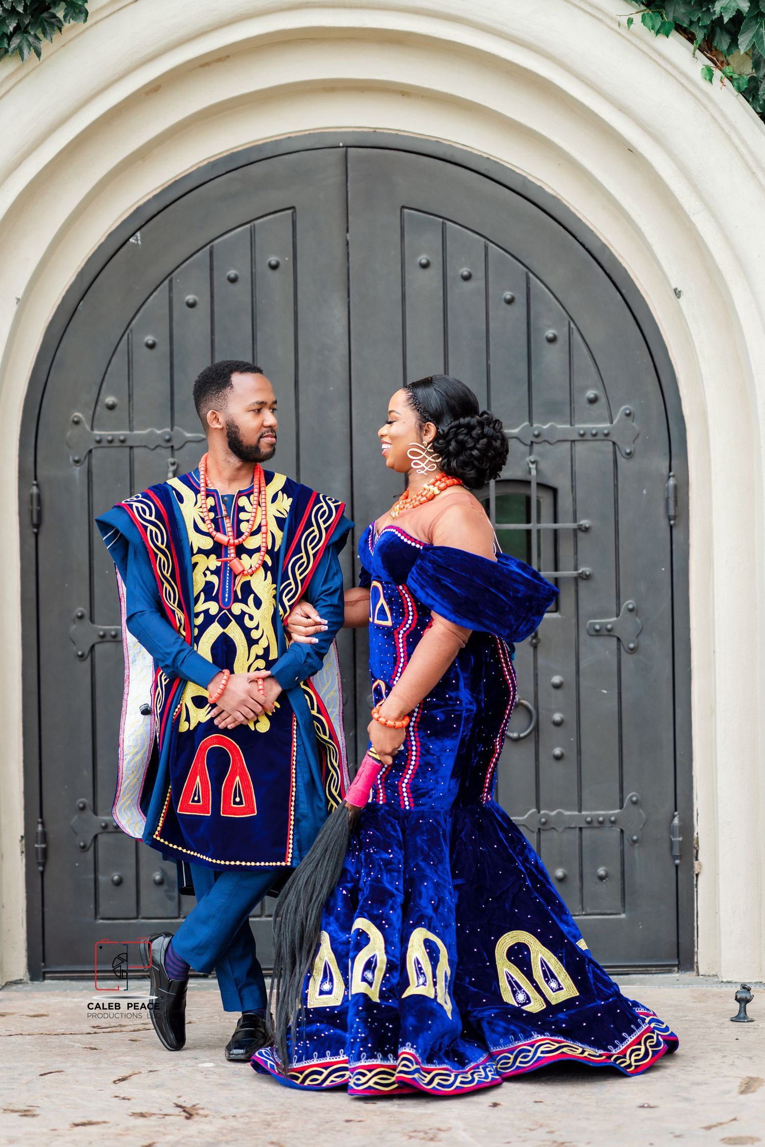 A couple in royal blue traditional African formal wear poses in front of an ornate arched doorway.