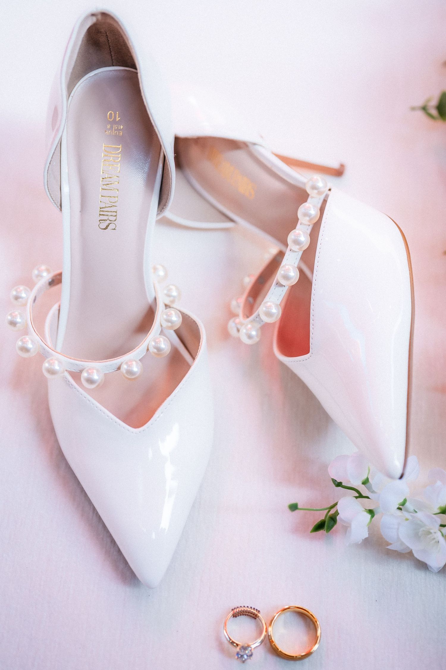 Elegant white patent leather bridal shoes and gold wedding rings arranged on soft pink background.