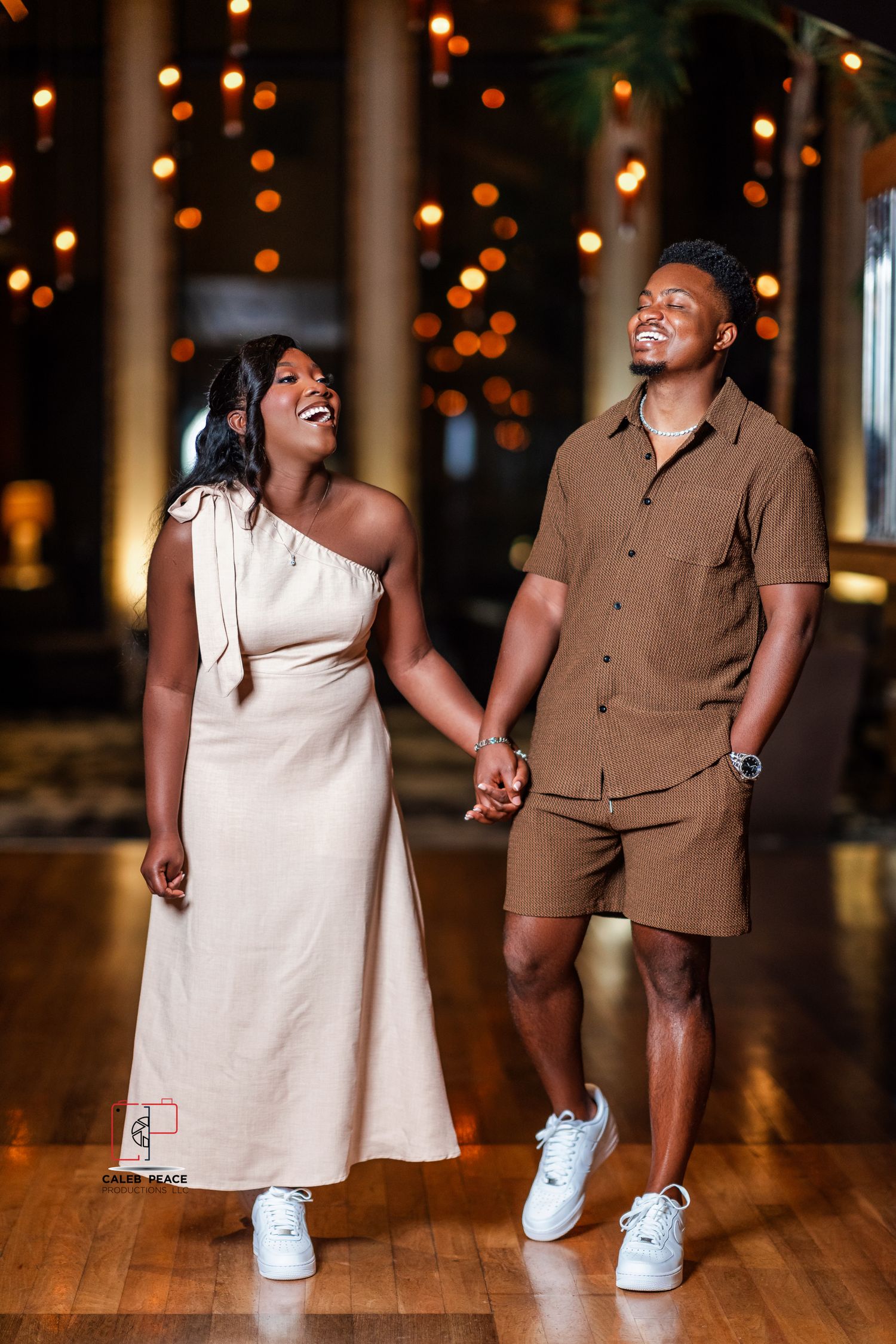 A couple in coordinating brown and white outfits dance playfully on a wooden floor with bokeh lights in the background.