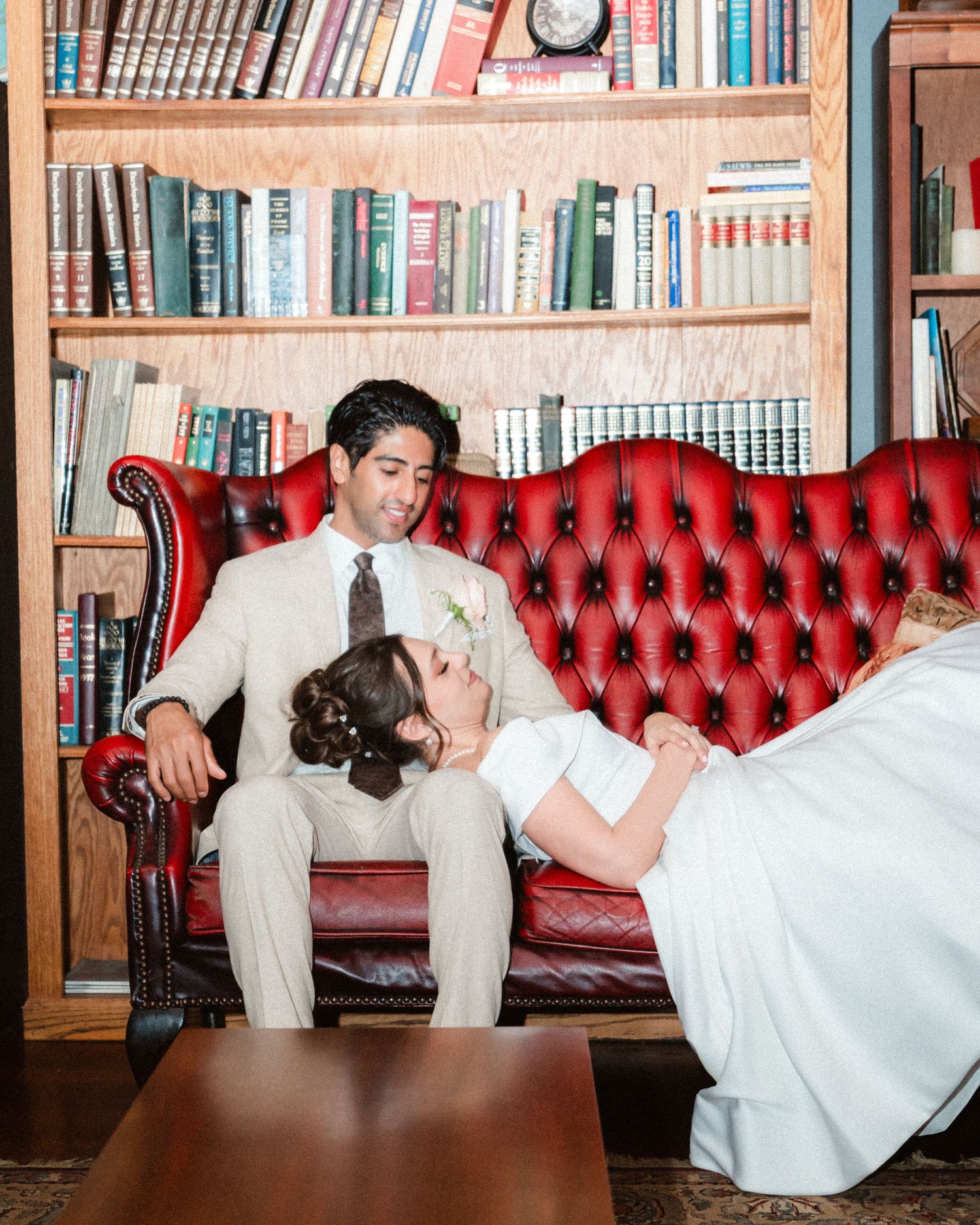 A couple in formal wear relaxes on a red leather couch in a library setting.