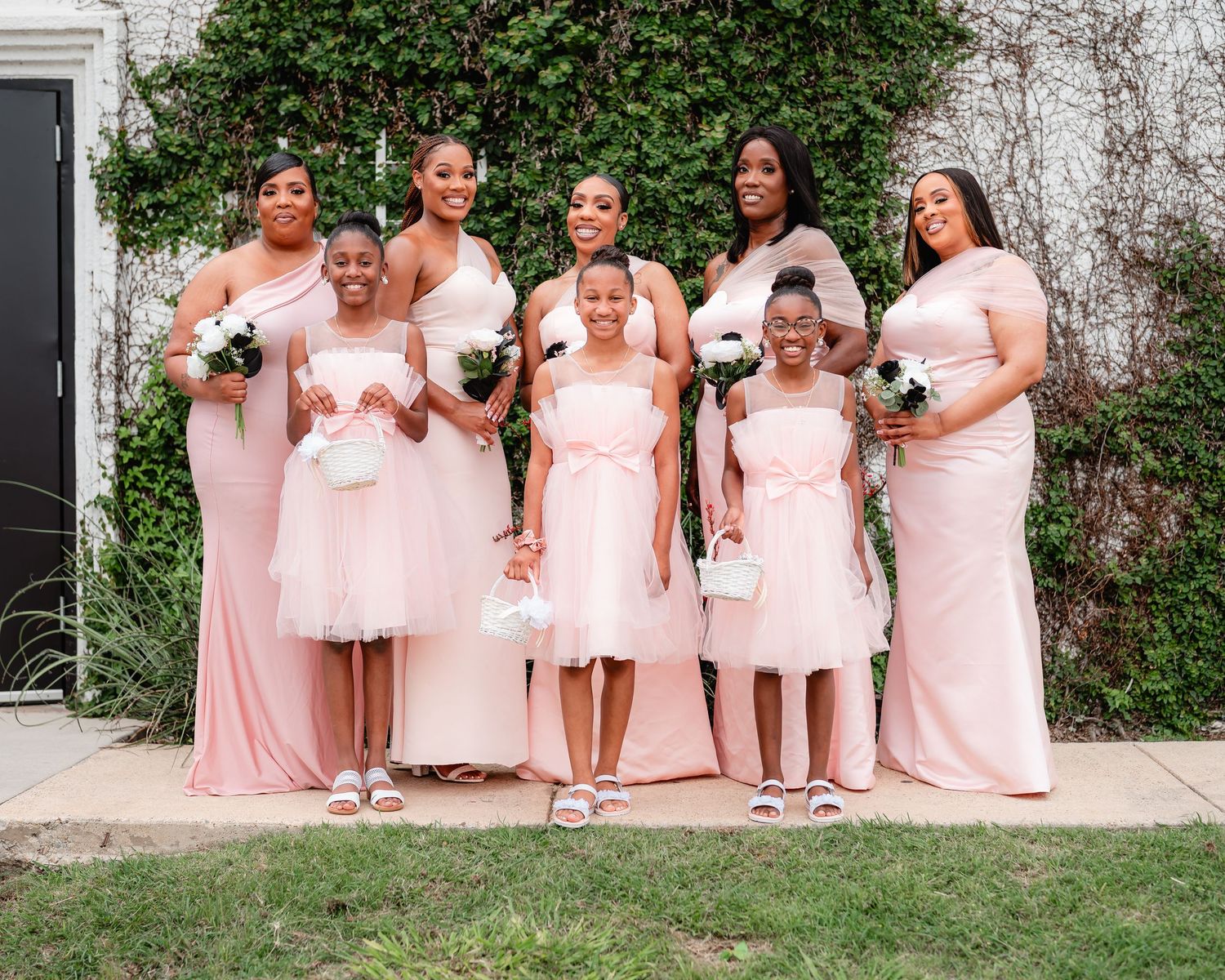 Bridesmaids in soft pink dresses lined up against a white wall with green foliage.