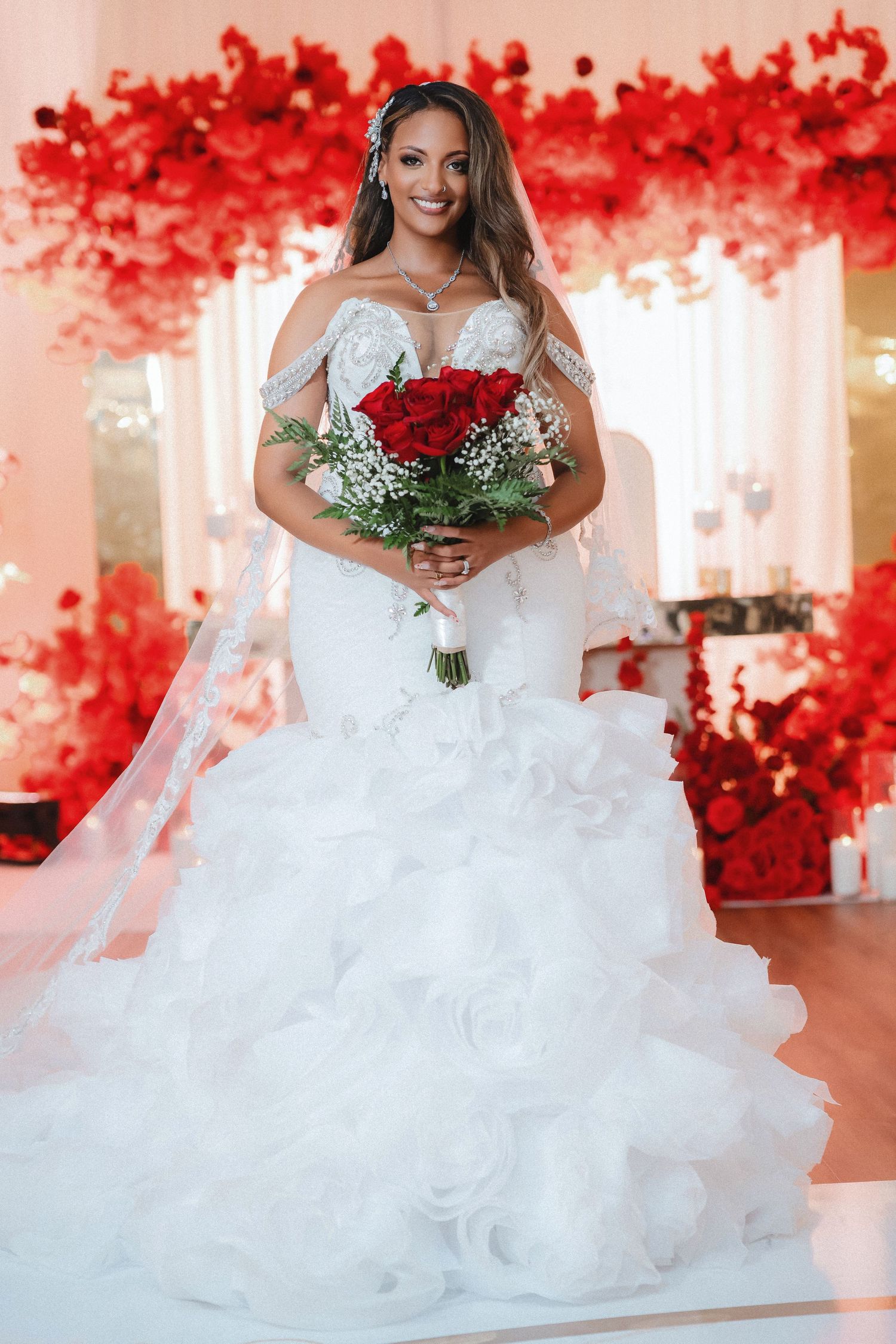 A bride in an elegant white off-shoulder gown holds a vibrant red rose bouquet against a backdrop of red floral decorations.