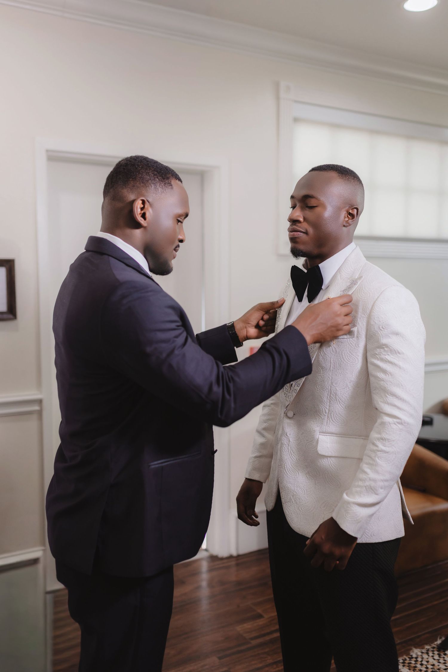 Two men in formal attire help adjust a white suit jacket and tie before a special occasion.