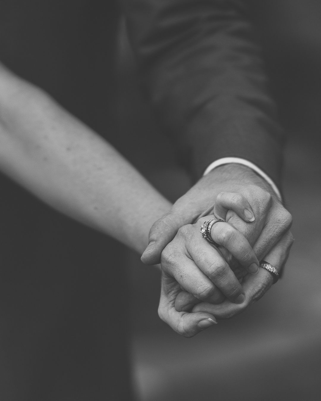 Black and white close-up of hands holding each other in a tender embrace, with wedding rings visible.