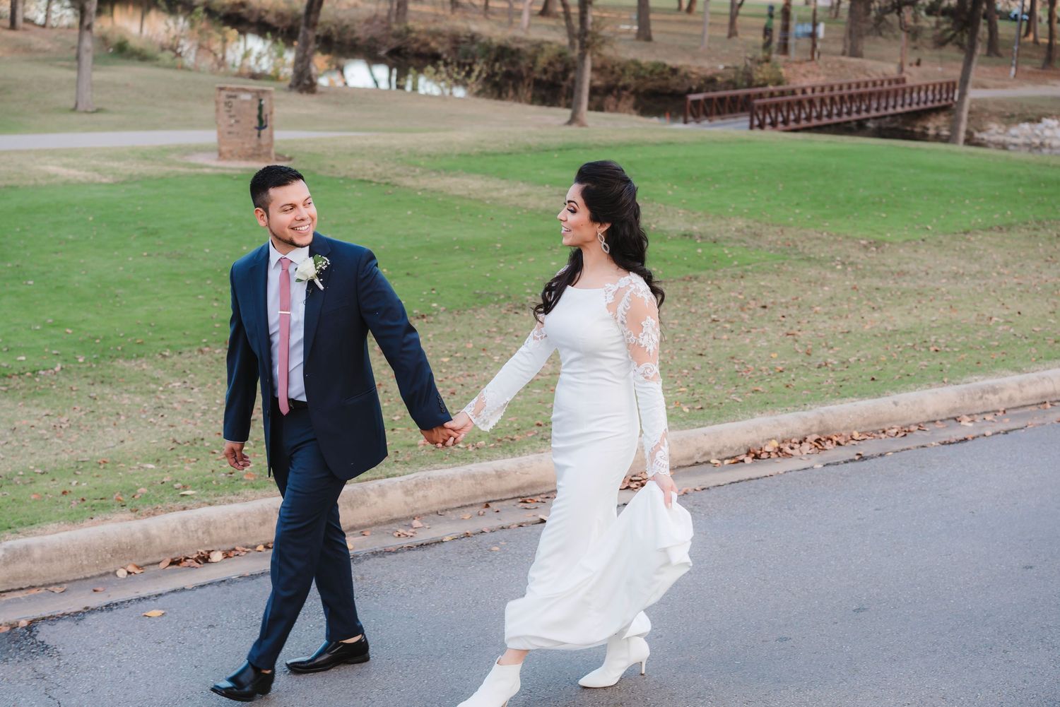 A couple in wedding attire playfully runs and dances together on a paved path surrounded by green lawns.
