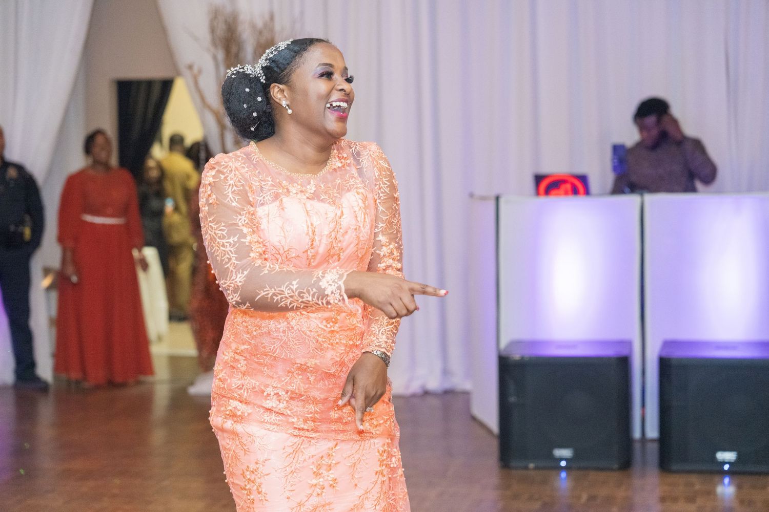 A guest dances at a wedding reception wearing a peach lace dress with DJ booth visible in background.