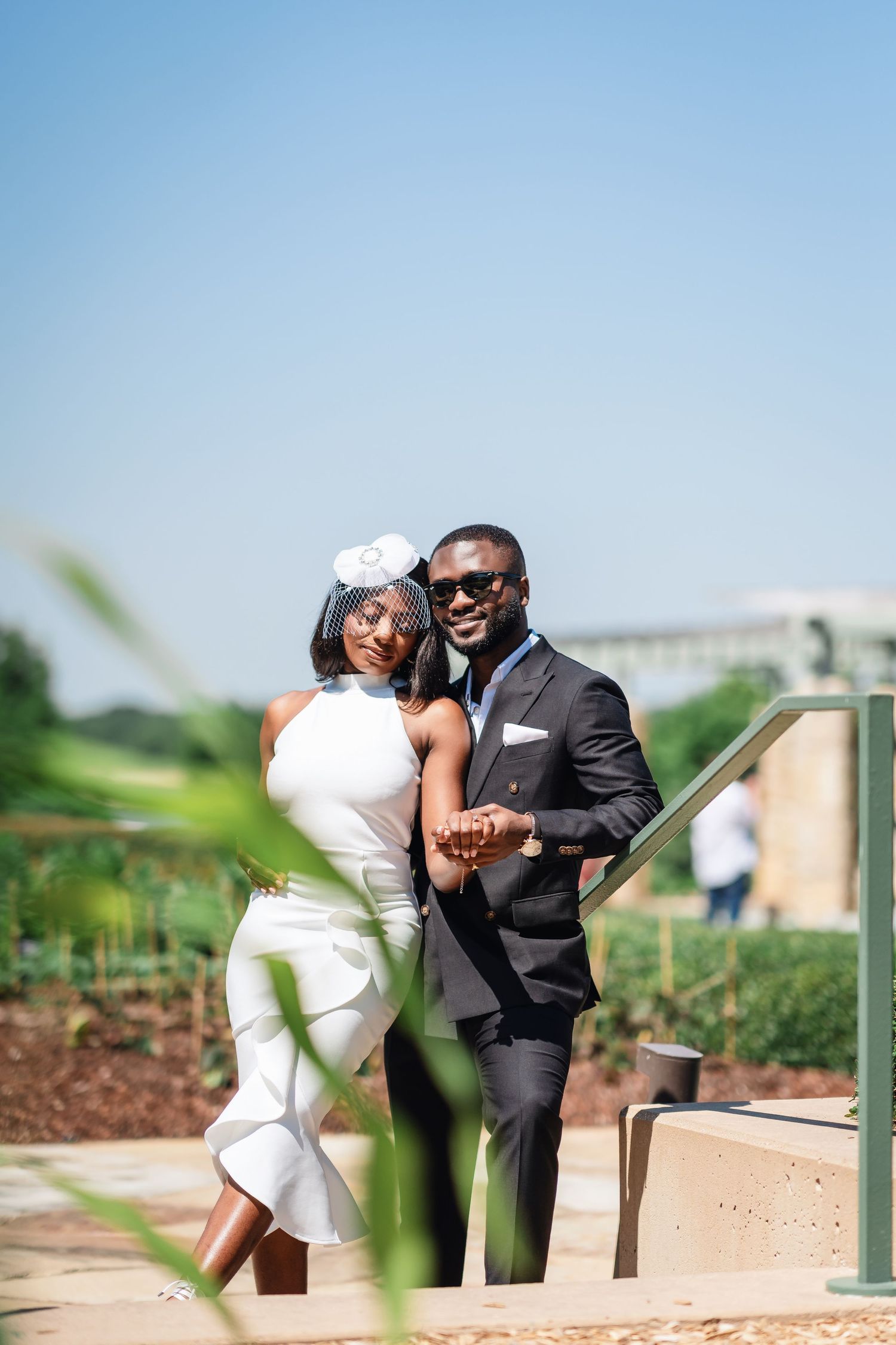 A couple in formal attire poses outdoors on a sunny day with the bride wearing a white dress and groom in dark suit.