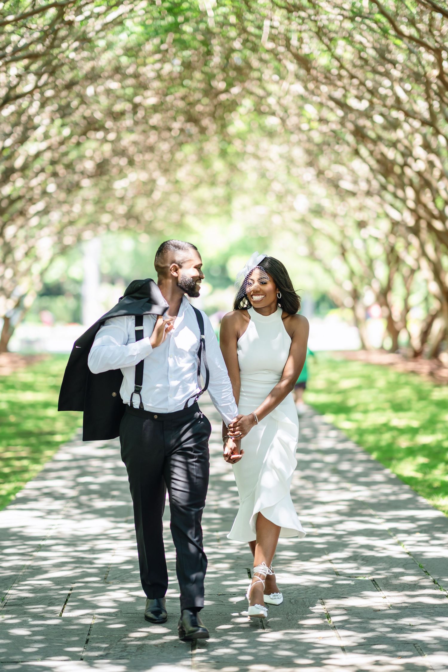 A joyful couple walks hand in hand down a tree-lined path in wedding attire.
