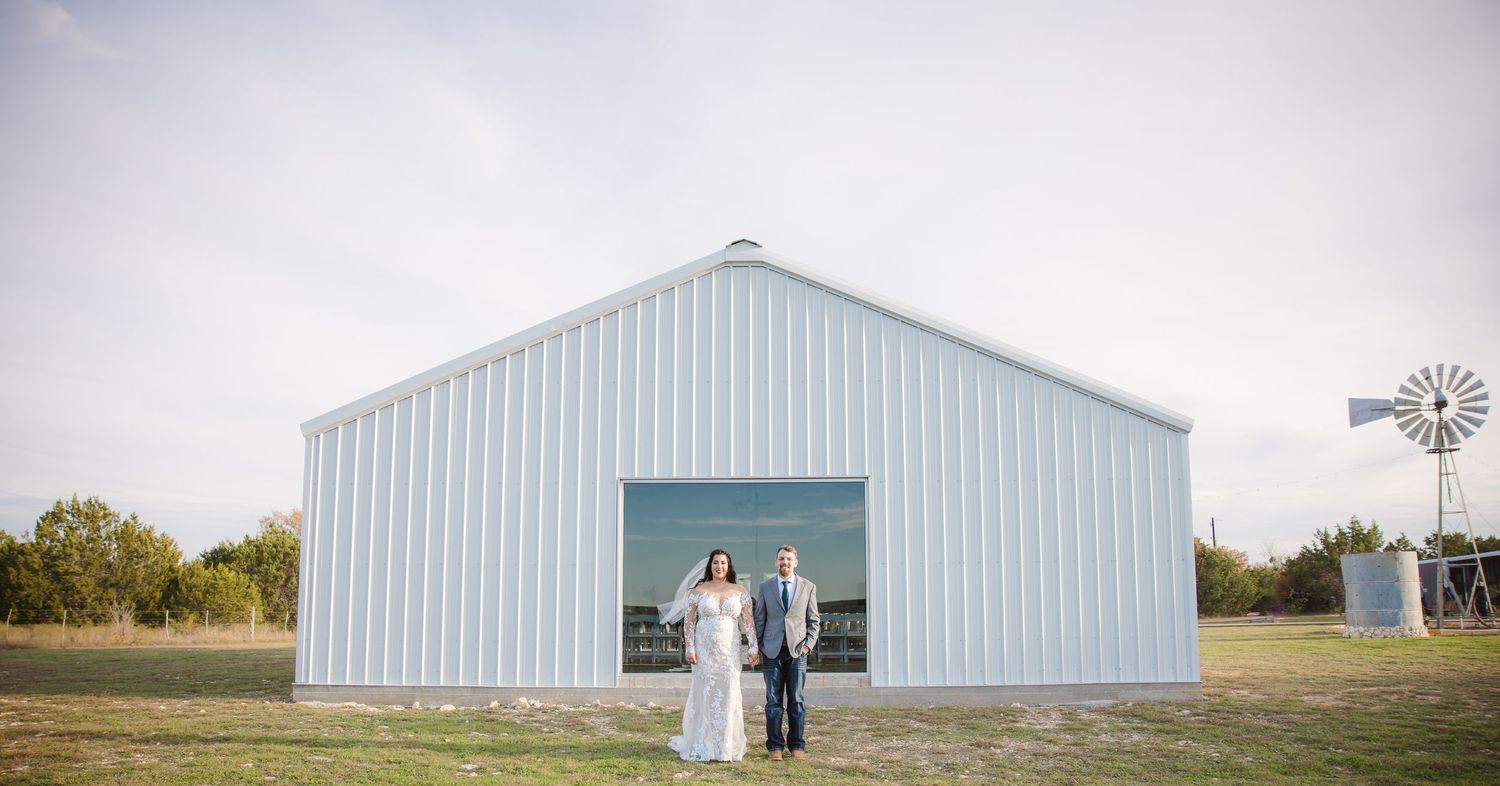 A couple stands in front of a white barn building during golden hour.