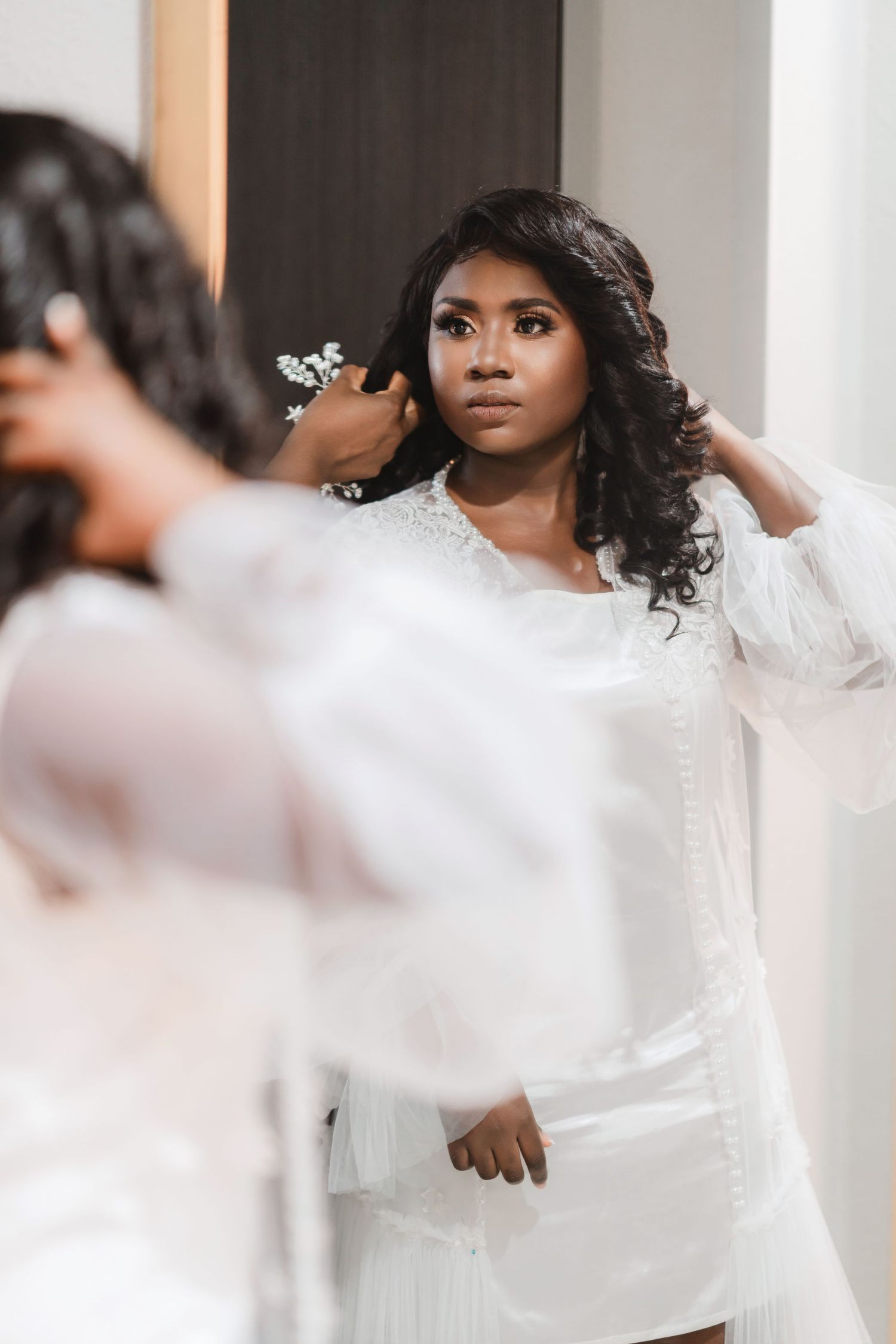 A bride in a white robe adjusts her curled hair while looking in a mirror during wedding preparations.