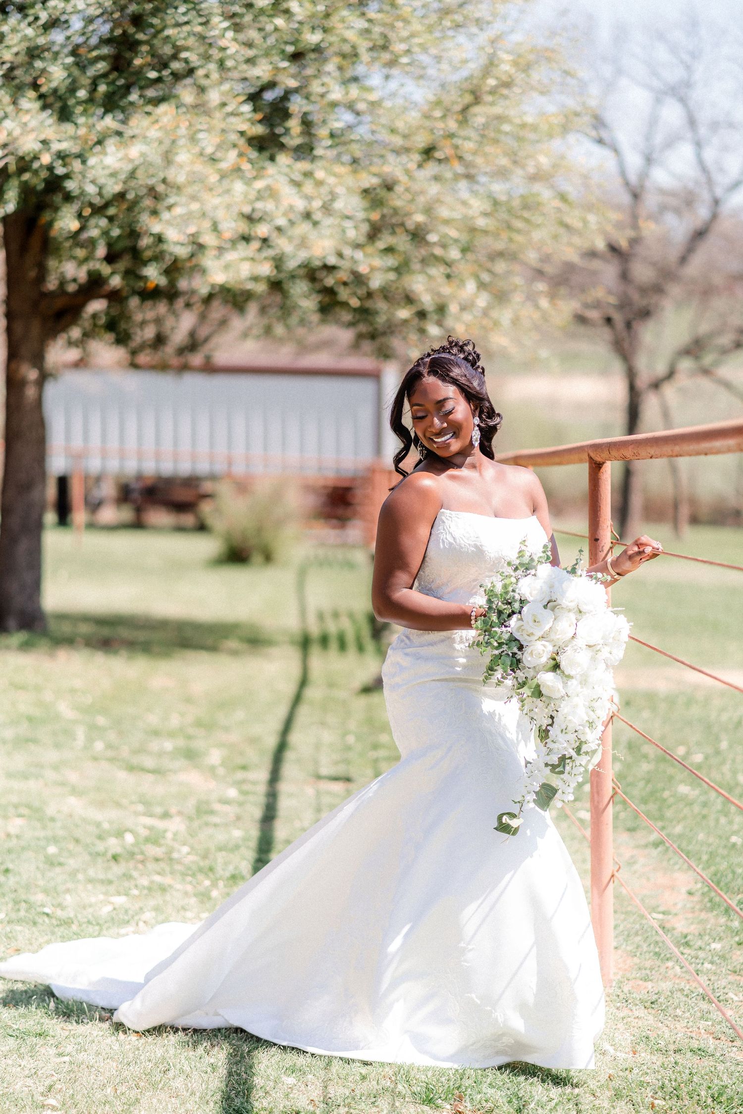 Bride in fitted white wedding gown poses outdoors holding cascading white bouquet against natural backdrop.