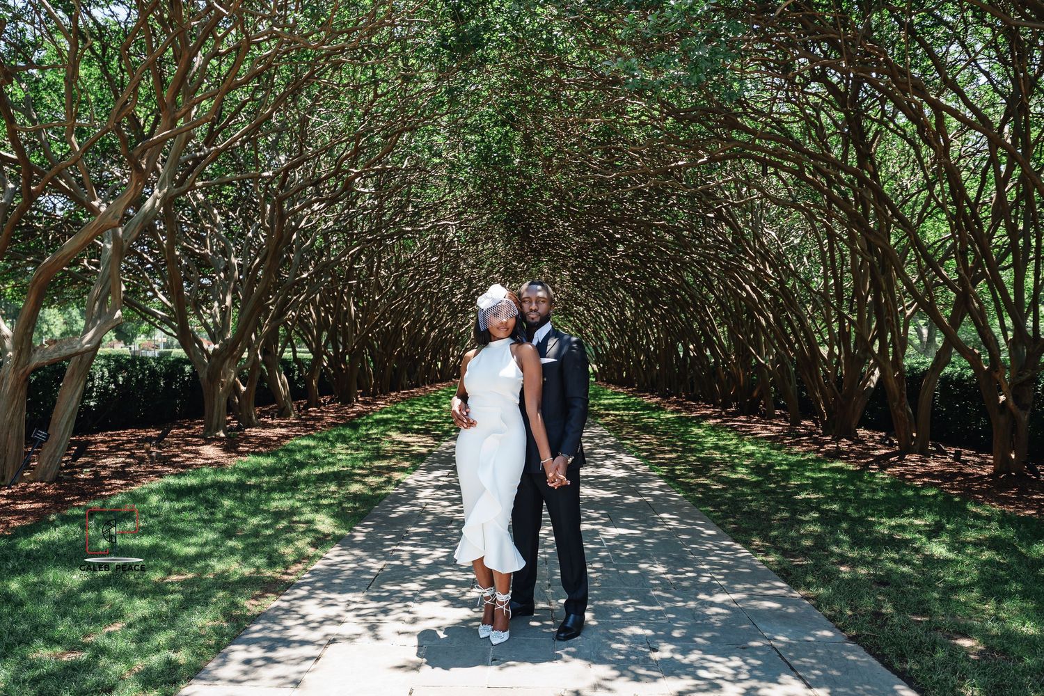 A couple stands together on a garden path beneath an arched canopy of lush green trees and branches.
