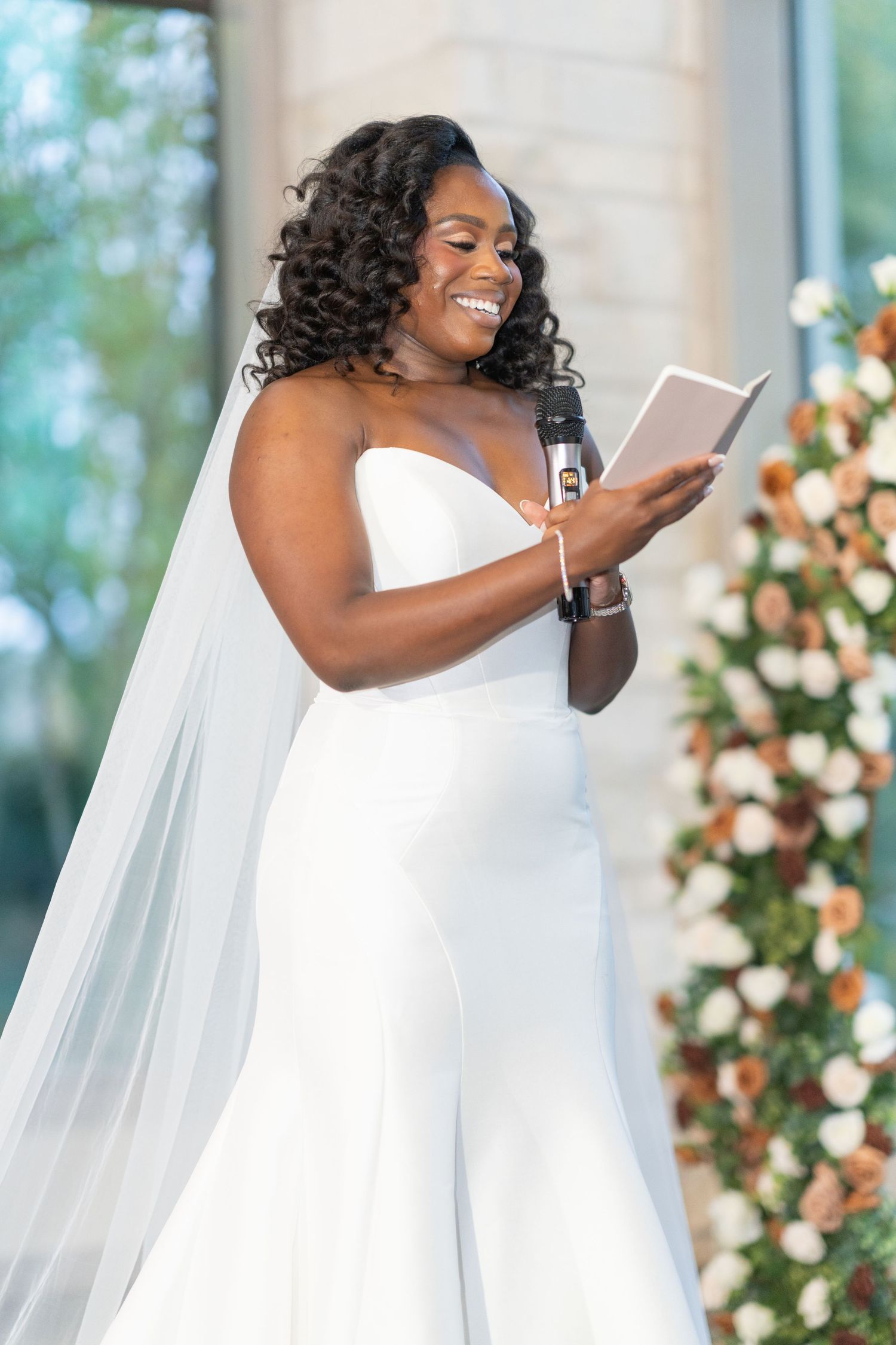 A bride in a flowing white dress and veil laughs while reading during the wedding ceremony.