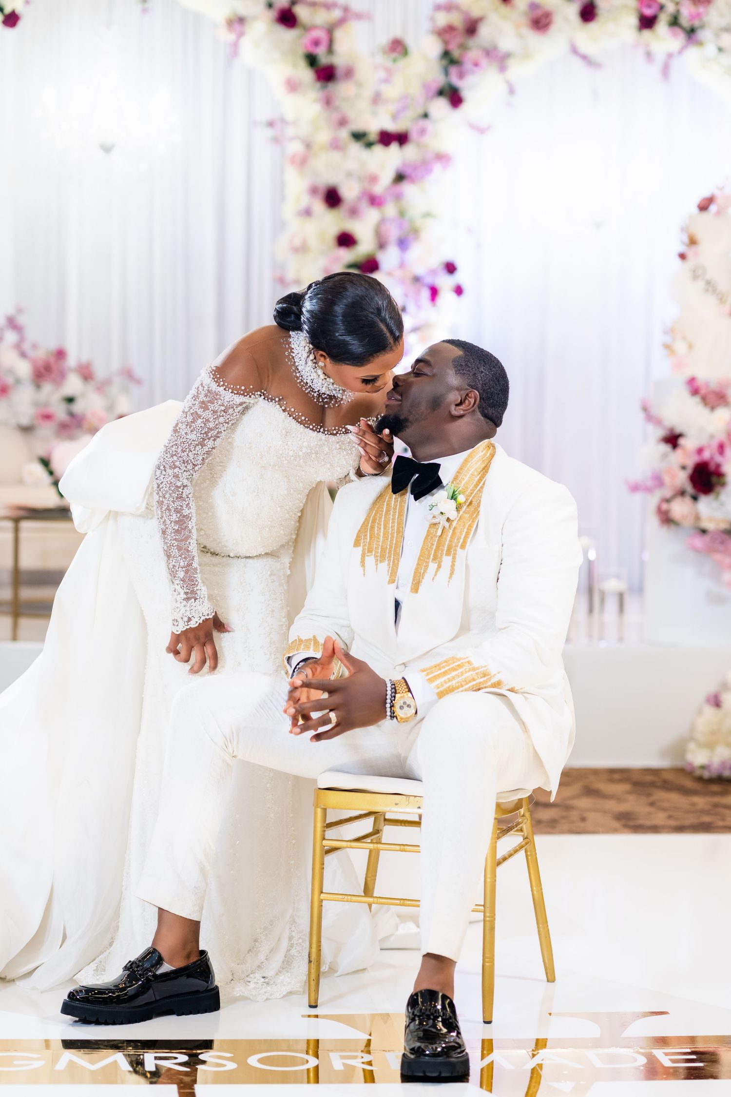 Couple in white attire sharing an intimate moment on gold chairs against a floral backdrop.